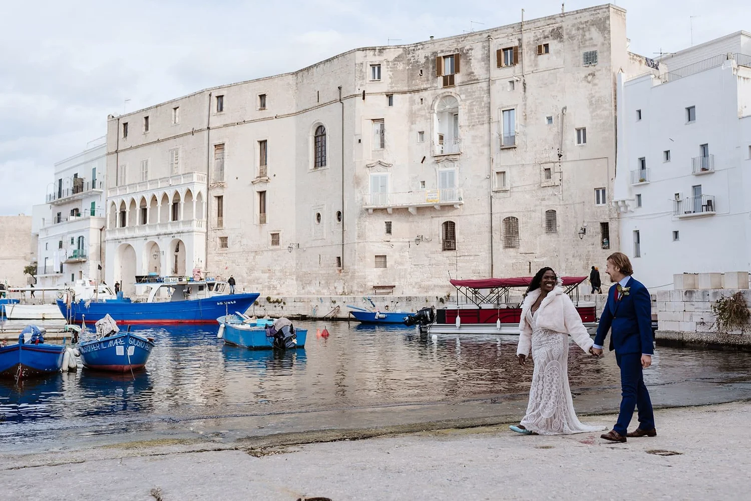 A newlywed couple holding hands walking along the waterfront in a European city, with boats docked in the water and old historic buildings in the background.
