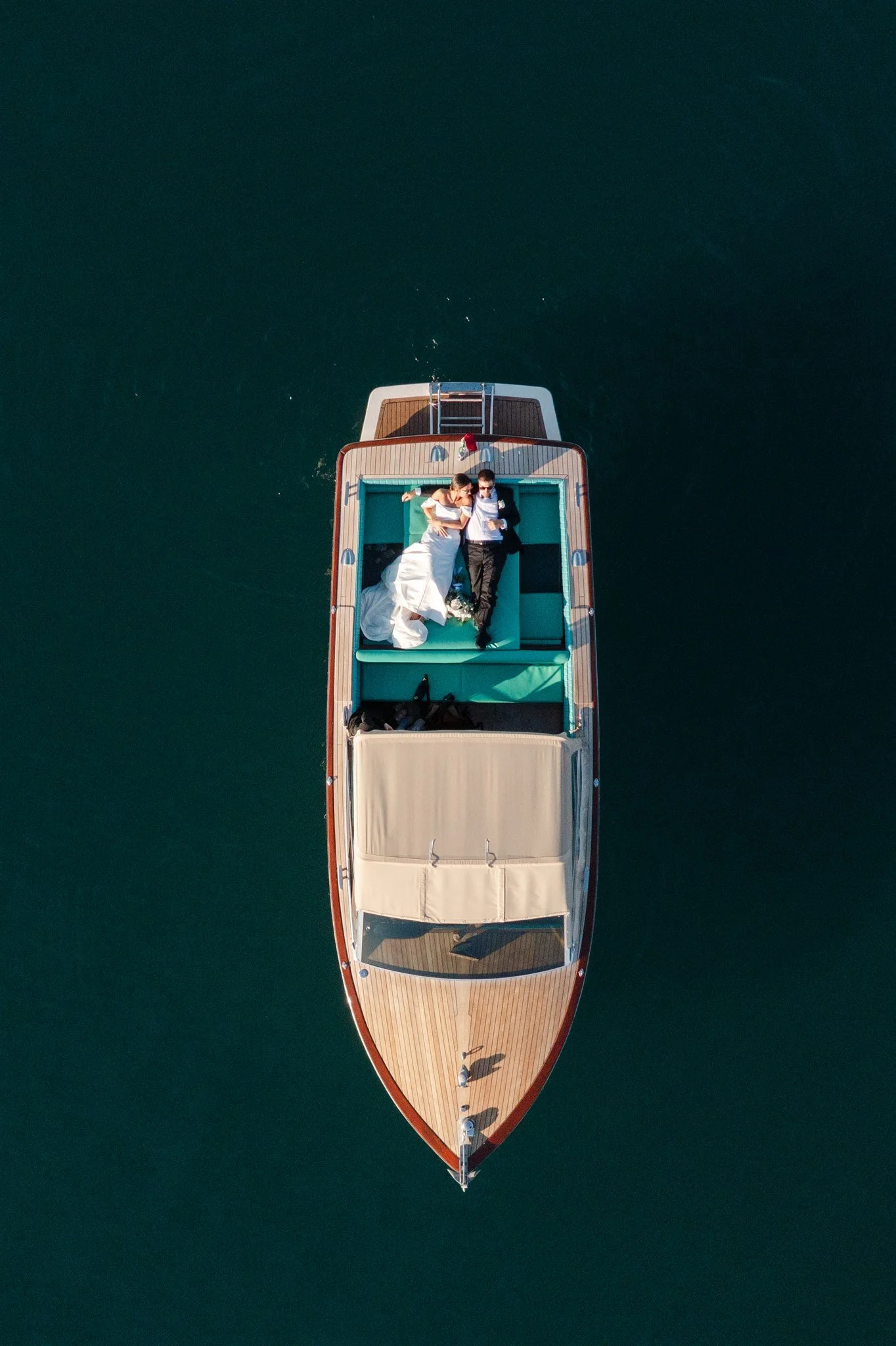 An aerial view of a boat with a bride and groom lying on its deck, floating on calm water.