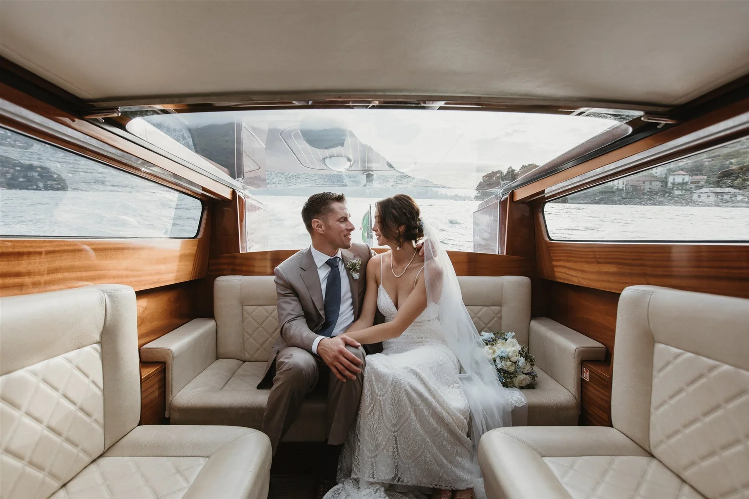 A bride and groom sitting closely inside a boat with wooden interior, water and a shoreline visible through the windows.