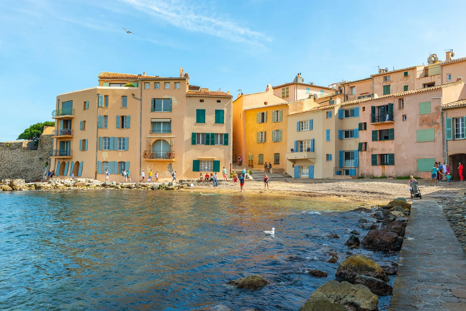 Colorful seaside buildings with green and blue shutters along a rocky shore, with people walking and ducks swimming in the water under a clear blue sky.