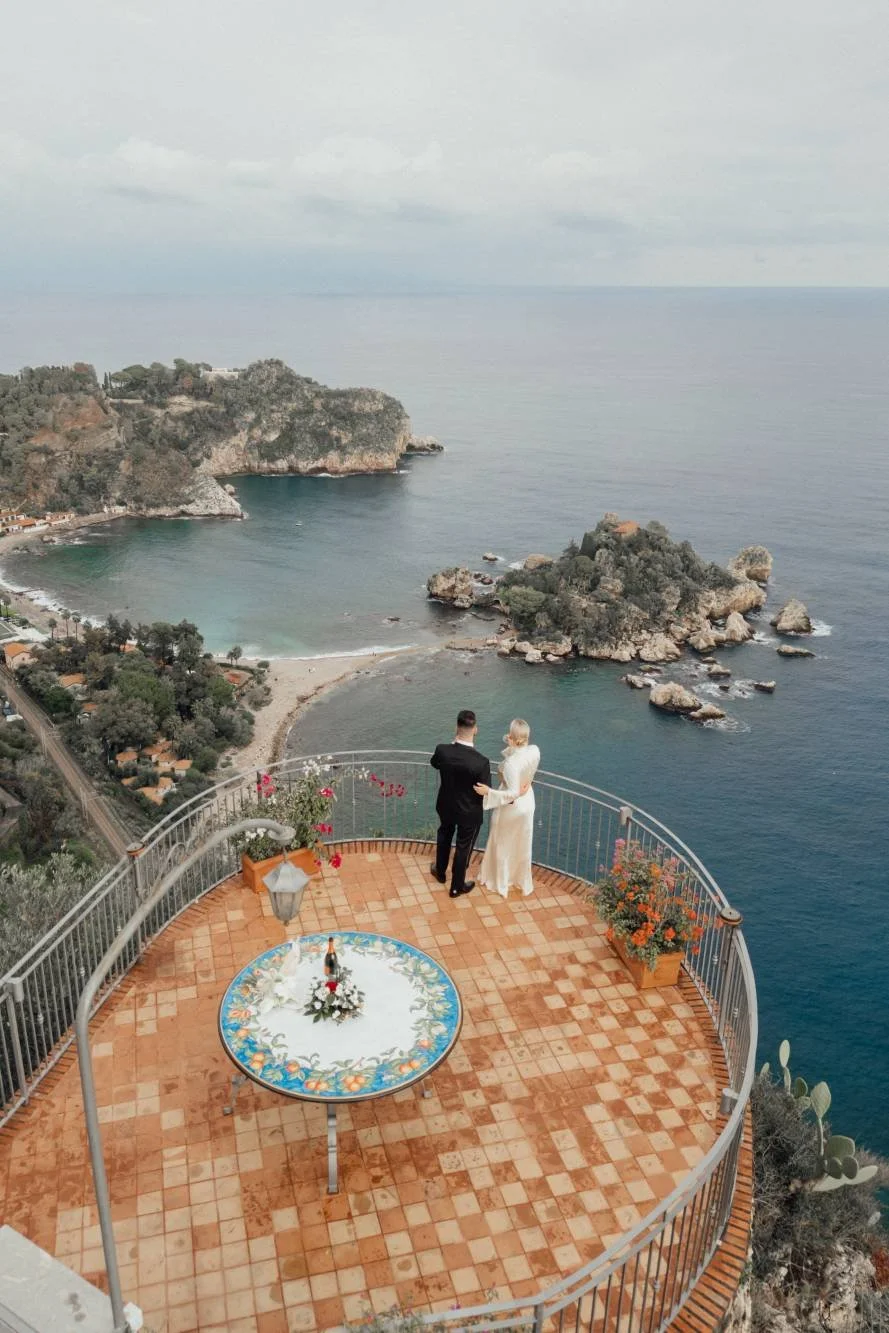 A couple dressed in wedding attire standing on a balcony overlooking a scenic coastal landscape with cliffs, water, and a small beach.