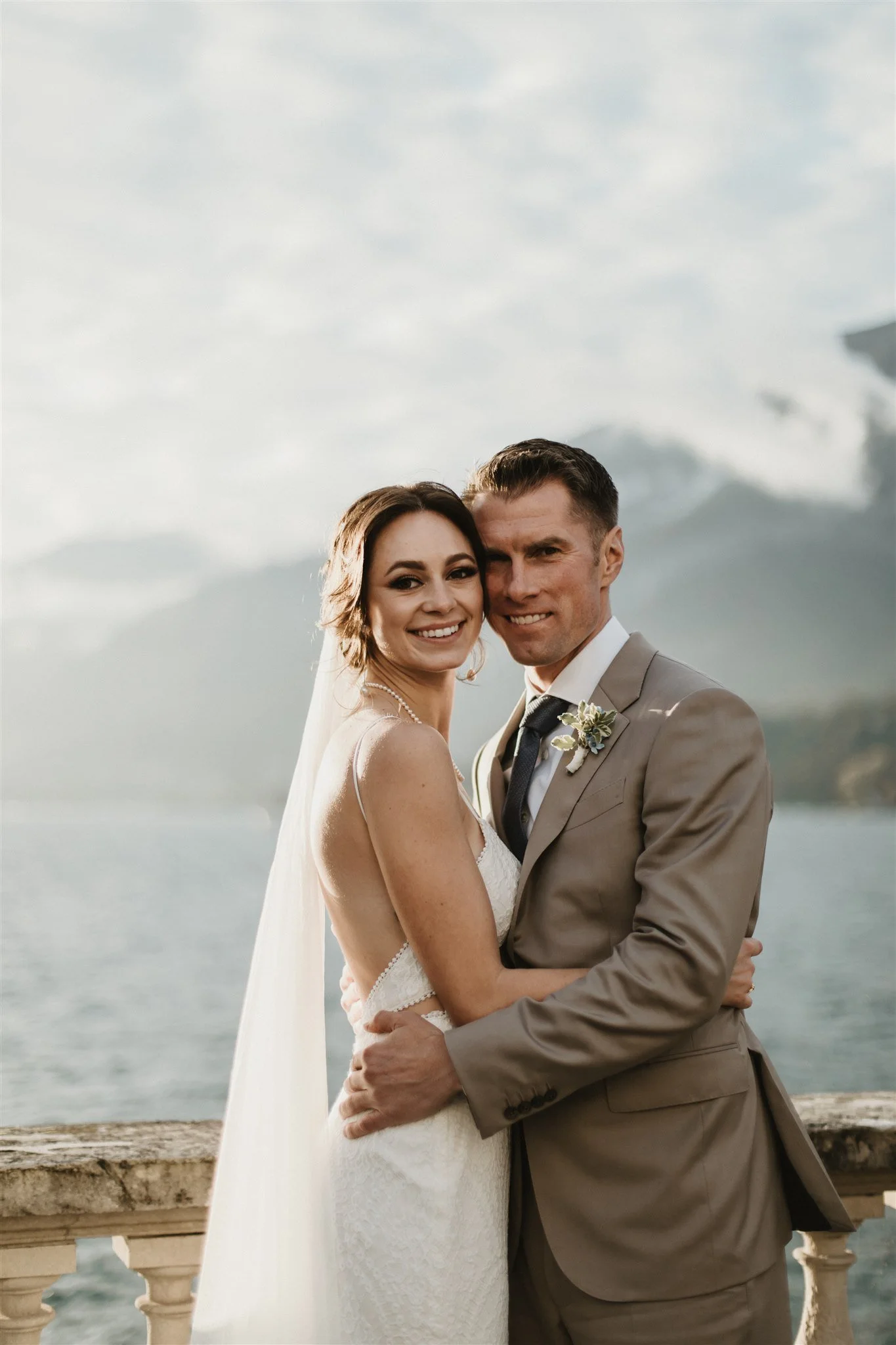 A smiling bride and groom embracing in front of a large body of water and a cloudy sky, during their wedding together.