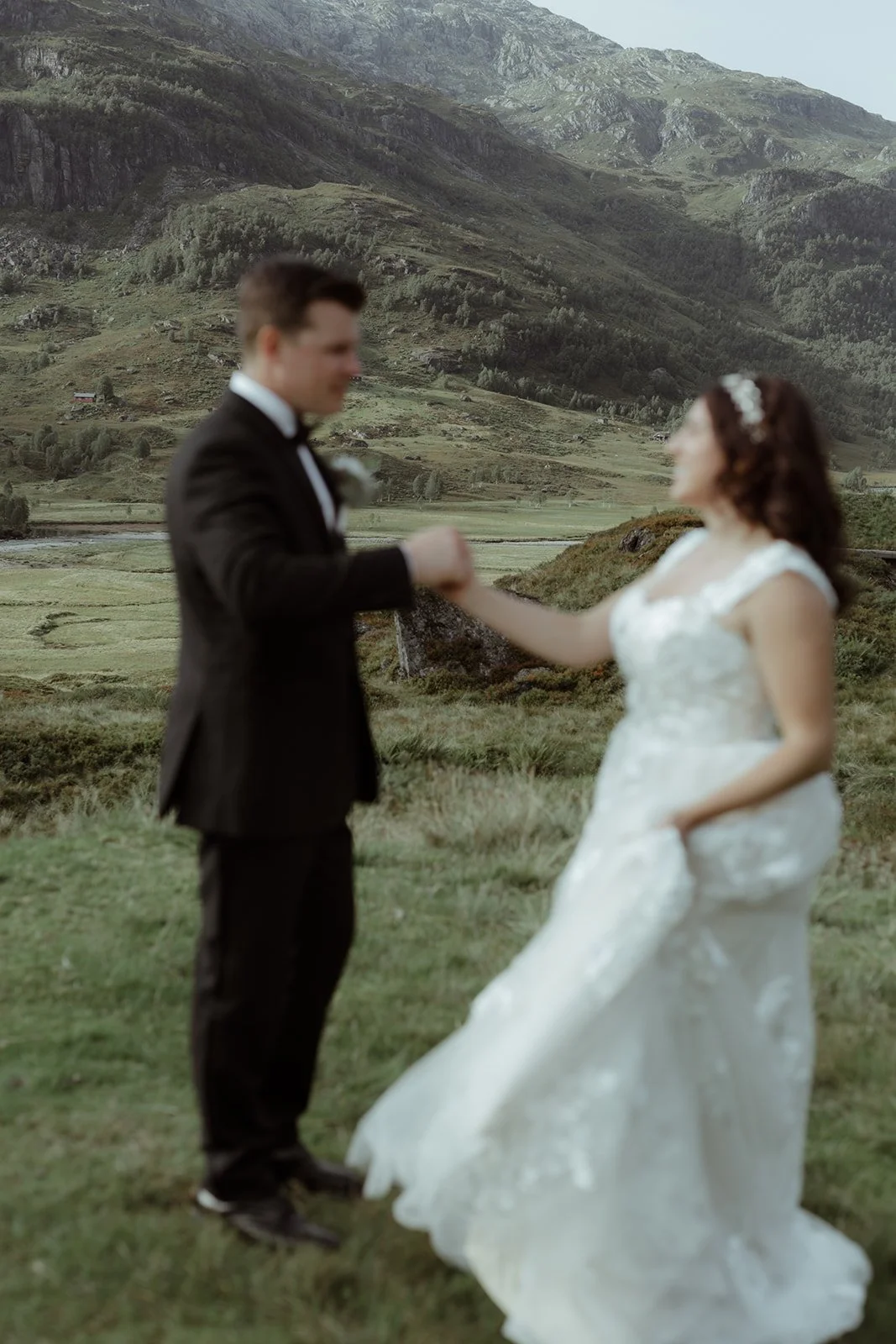 A bride and groom holding hands in a lush green field with mountains in the background, during a wedding photoshoot.