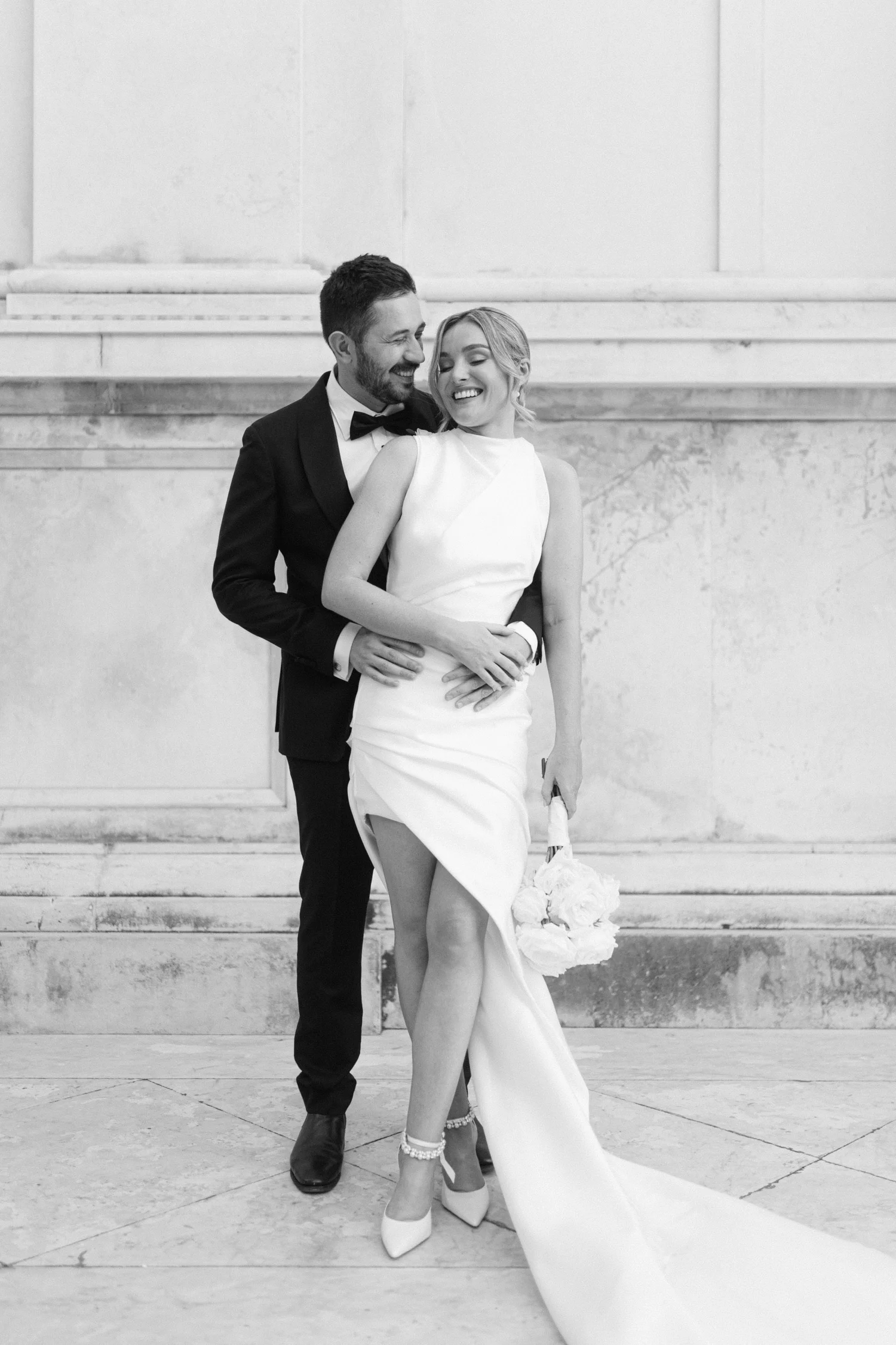 A black-and-white photograph of a smiling couple in wedding attire, standing on steps in front of a stone wall. The groom is wearing a tuxedo with a bow tie, with his arm around the bride's waist. The bride is wearing a sleeveless white wedding dress