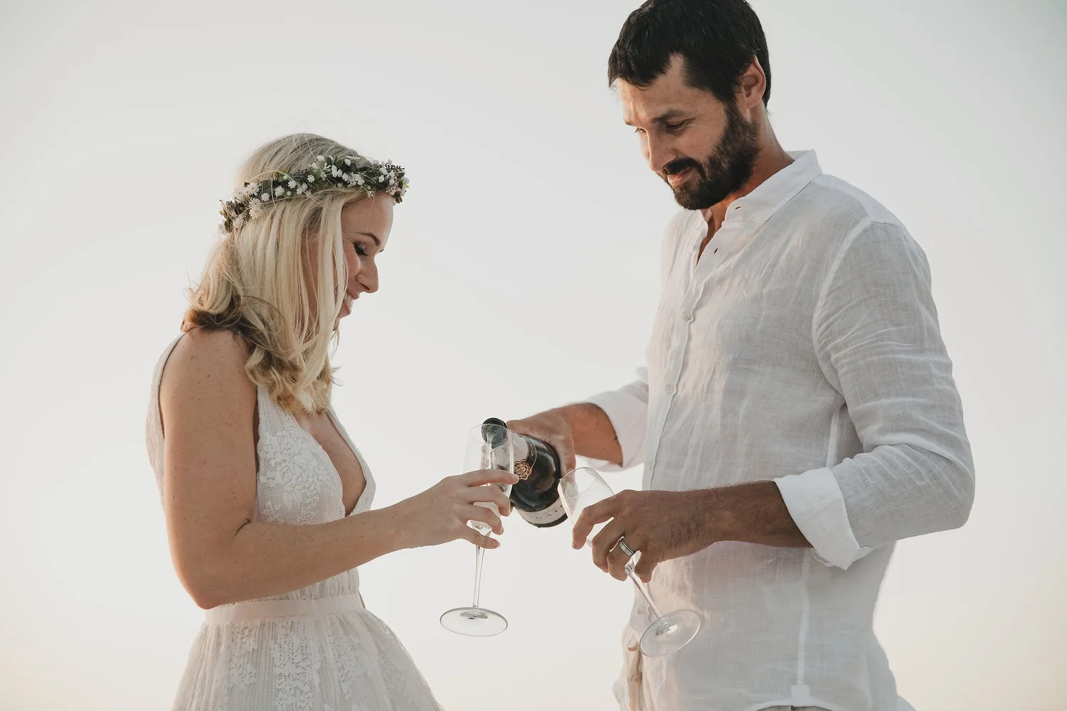 A man and woman are celebrating with glasses of wine; the woman wears a floral crown and white dress, and the man wears a white shirt, both smiling softly.