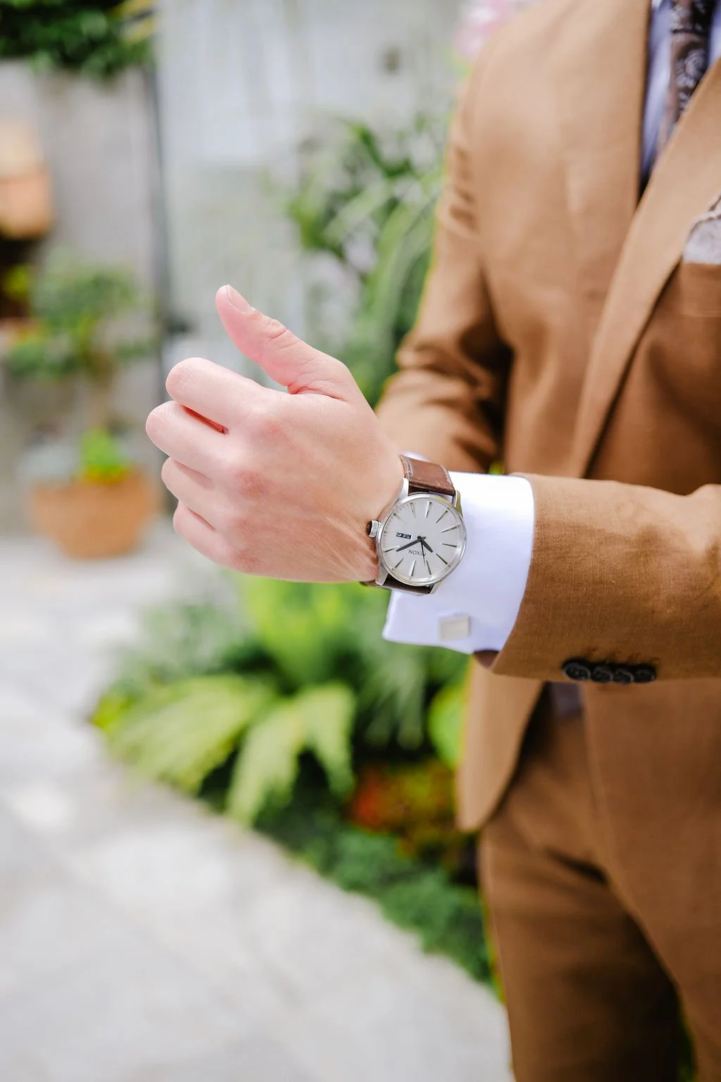 A person wearing a brown suit and white shirt looking at a wristwatch, standing in a green garden or greenhouse.