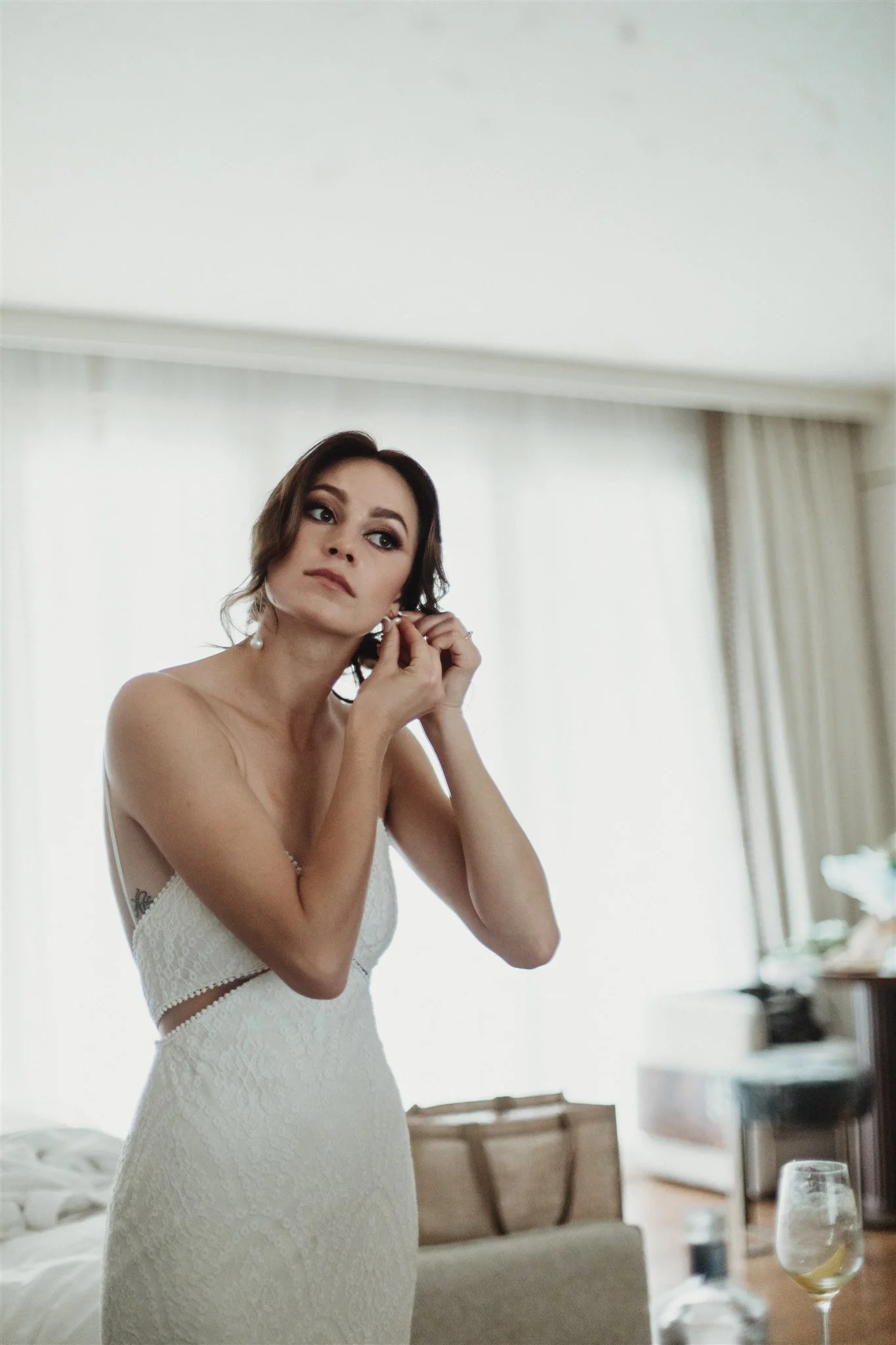 Woman in a white lace dress is putting on earrings in a room with natural light and neutral decor.