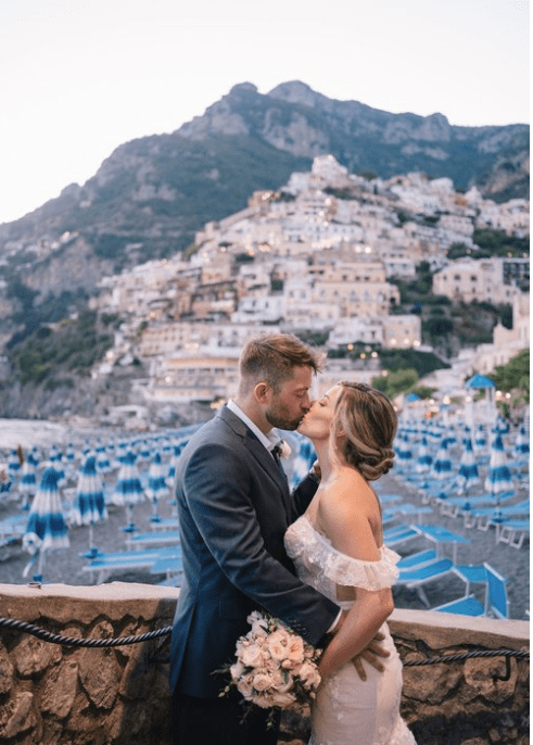 A couple in wedding attire sharing a kiss on a seaside promenade with a backdrop of a cliffside town and beach umbrellas.