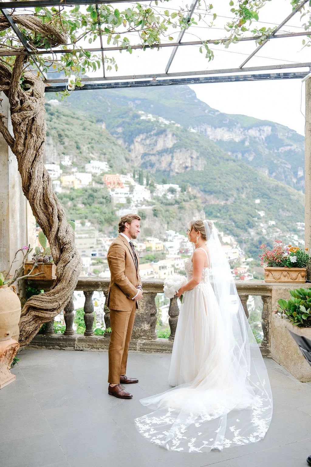 A bride and groom exchange vows on a terrace with scenic mountain views in Italy, surrounded by plants and flowers.