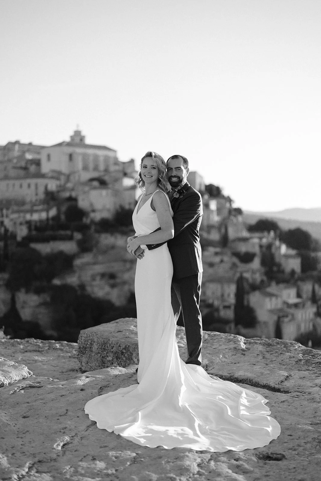 Black and white photo of a bride and groom standing outdoors on a rocky surface, with a historic cityscape in the background. The bride is in a long, elegant wedding dress, and the groom is in a suit, embracing her from behind with a smile.