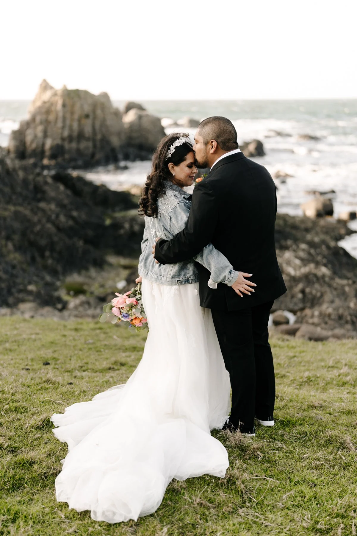 A bride and groom stand close together on a grassy area near the ocean, embracing. The bride is wearing a white wedding gown and a denim jacket, holding a bouquet of pink, purple, and white flowers. The groom is wearing a black suit and kissing the b
