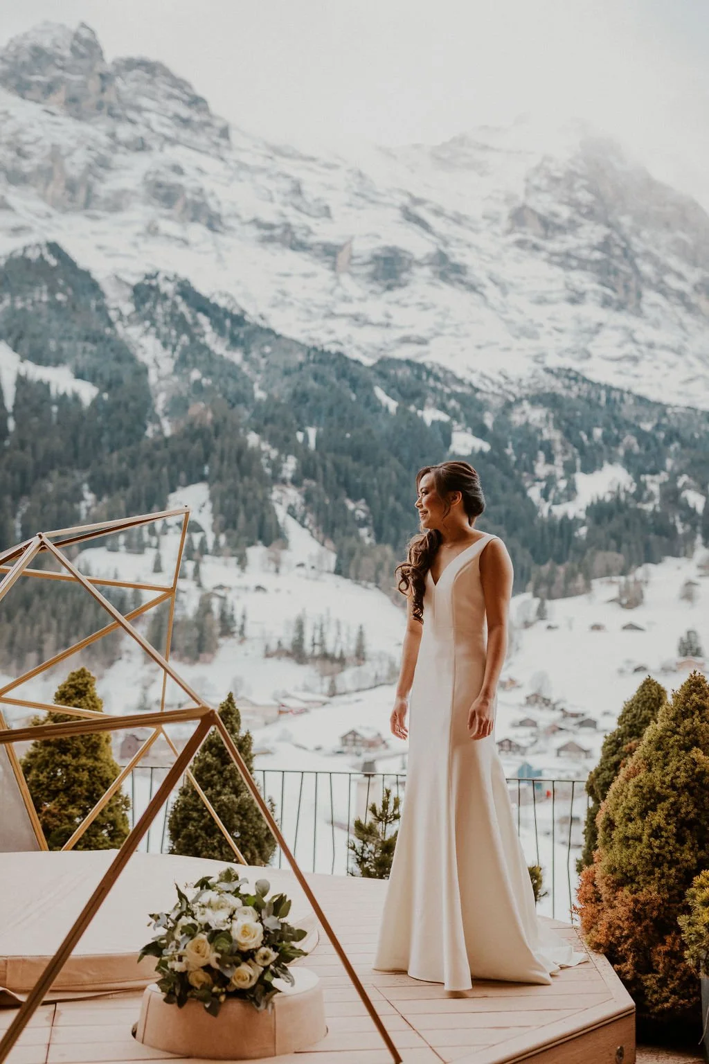 A bride in a white gown standing outdoors on a wooden platform with snow-covered mountains and trees in the background, surrounded by greenery and a floral arrangement in the foreground.