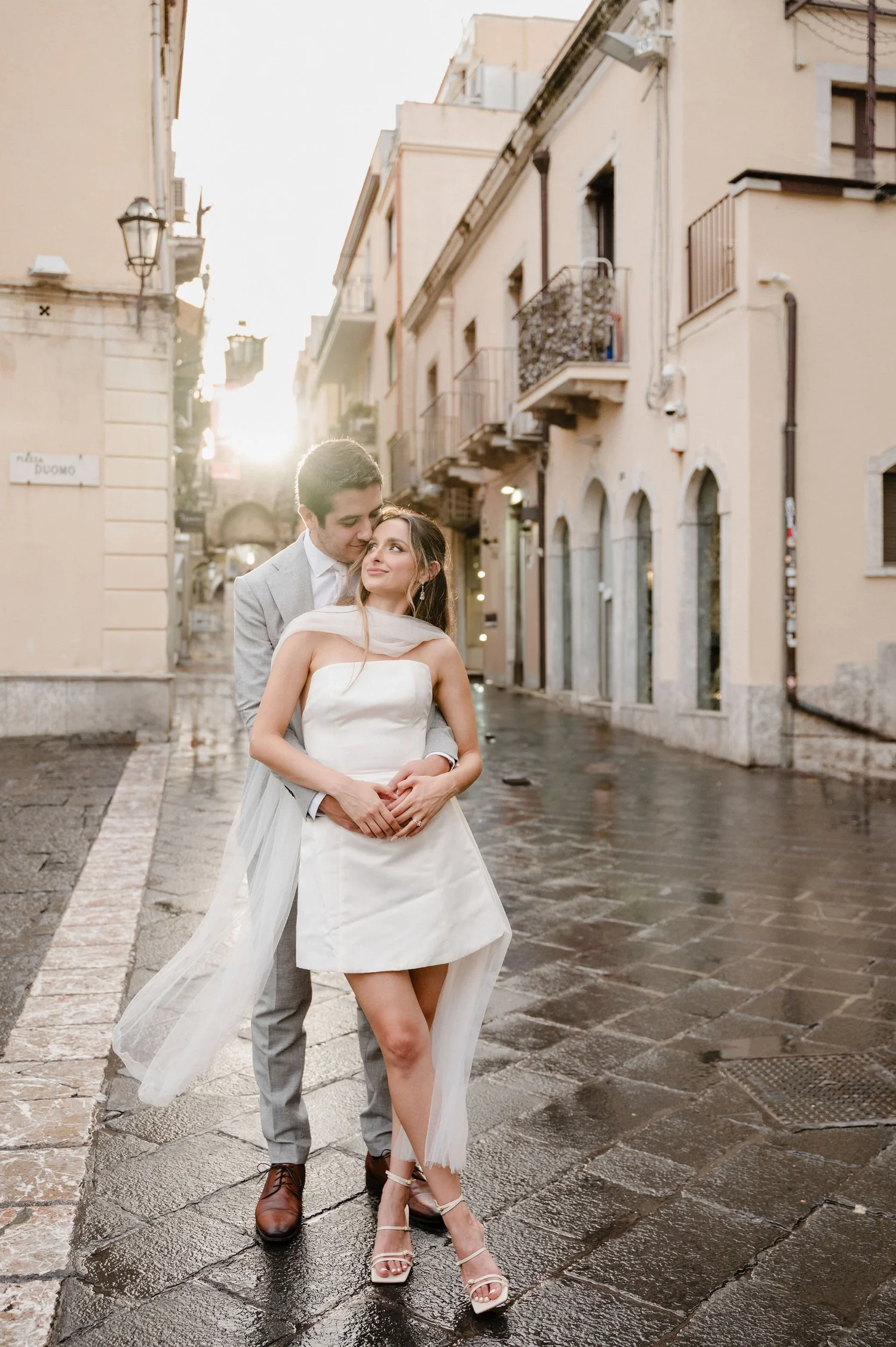 A bride and groom standing closely together on a wet city street during sunset, with narrow buildings and balconies in the background.