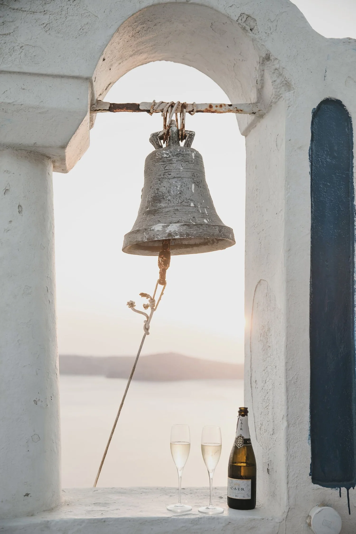A rustic stone bell hangs from an archway against a white wall, with two glasses of champagne and a bottle of champagne on the ledge below.