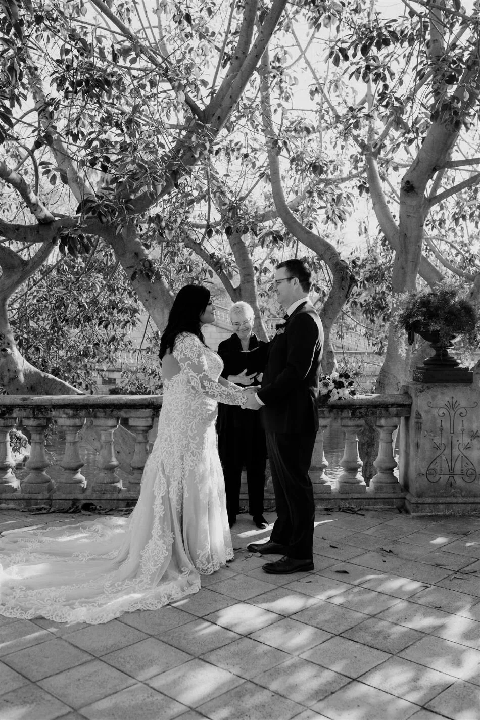 Black and white photo of a couple holding hands and exchanging vows during a wedding ceremony outdoors, with an officiant standing behind them and a large tree in the background.