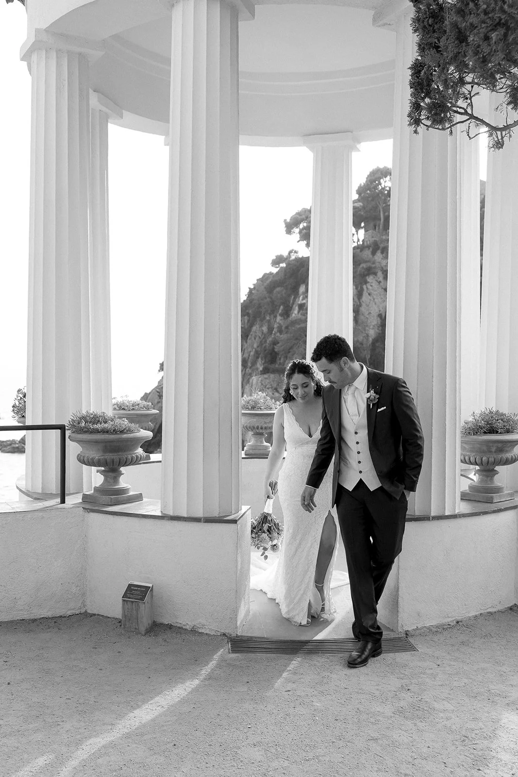A black and white photo of a newlywed couple walking out of a classical temple with tall columns, holding hands and smiling, with a scenic landscape in the background.