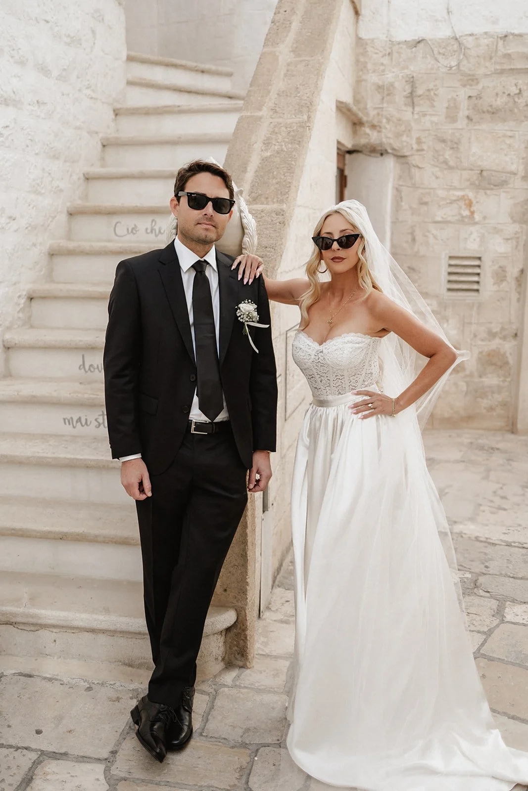 A bride and groom in wedding attire standing against a stone staircase and wall. The groom is wearing a black suit with a white shirt, black tie, and sunglasses. The bride is in a white strapless wedding gown with lace bodice, long flowy skirt, veil,