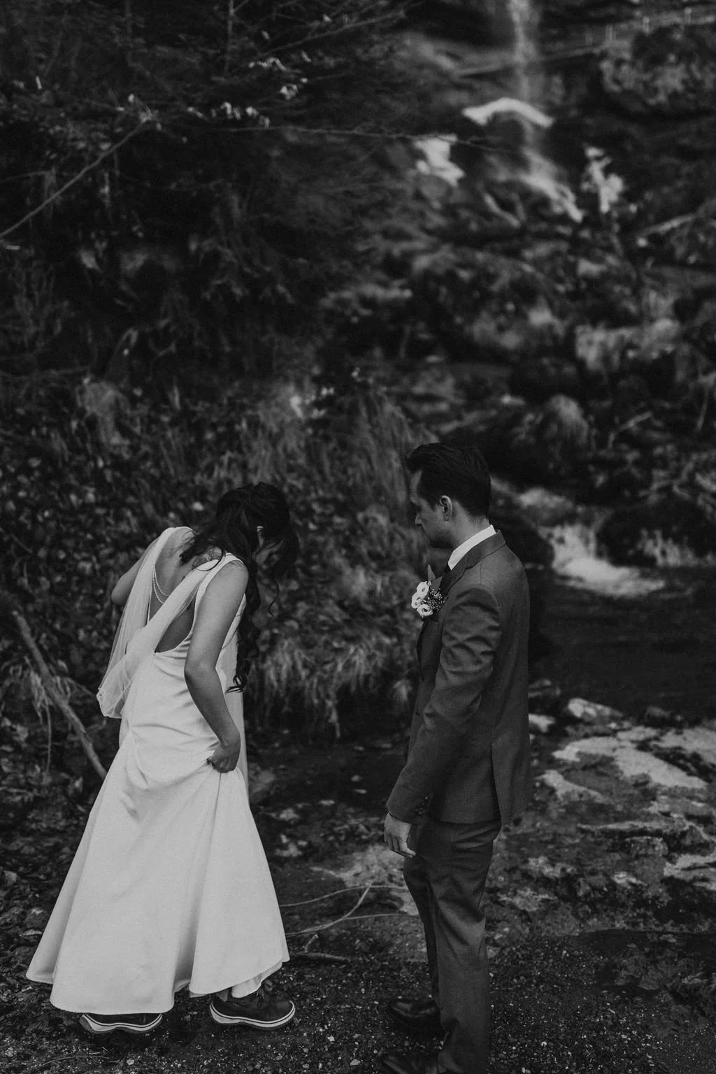 A black and white photo of a bride and groom standing near a rocky stream. The bride is wearing a long white wedding gown and the groom is dressed in a suit with a boutonniere.