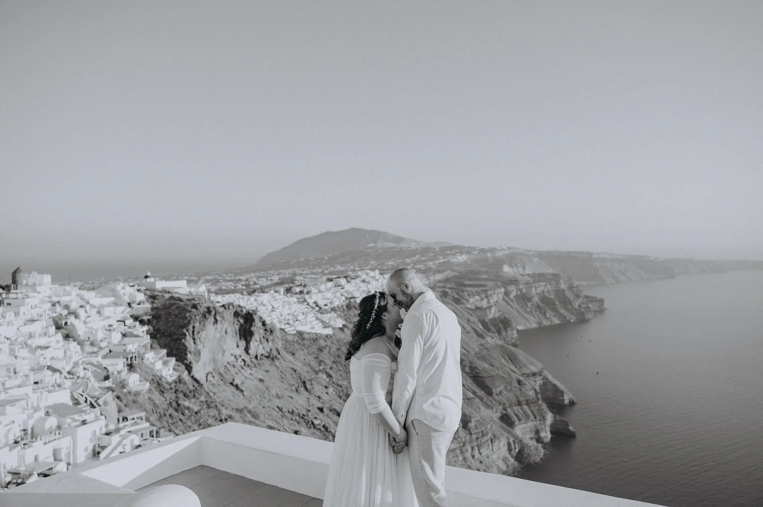 A couple in wedding attire sharing a kiss on a balcony overlooking a seaside landscape with hillside houses and a distant mountain, in black and white.