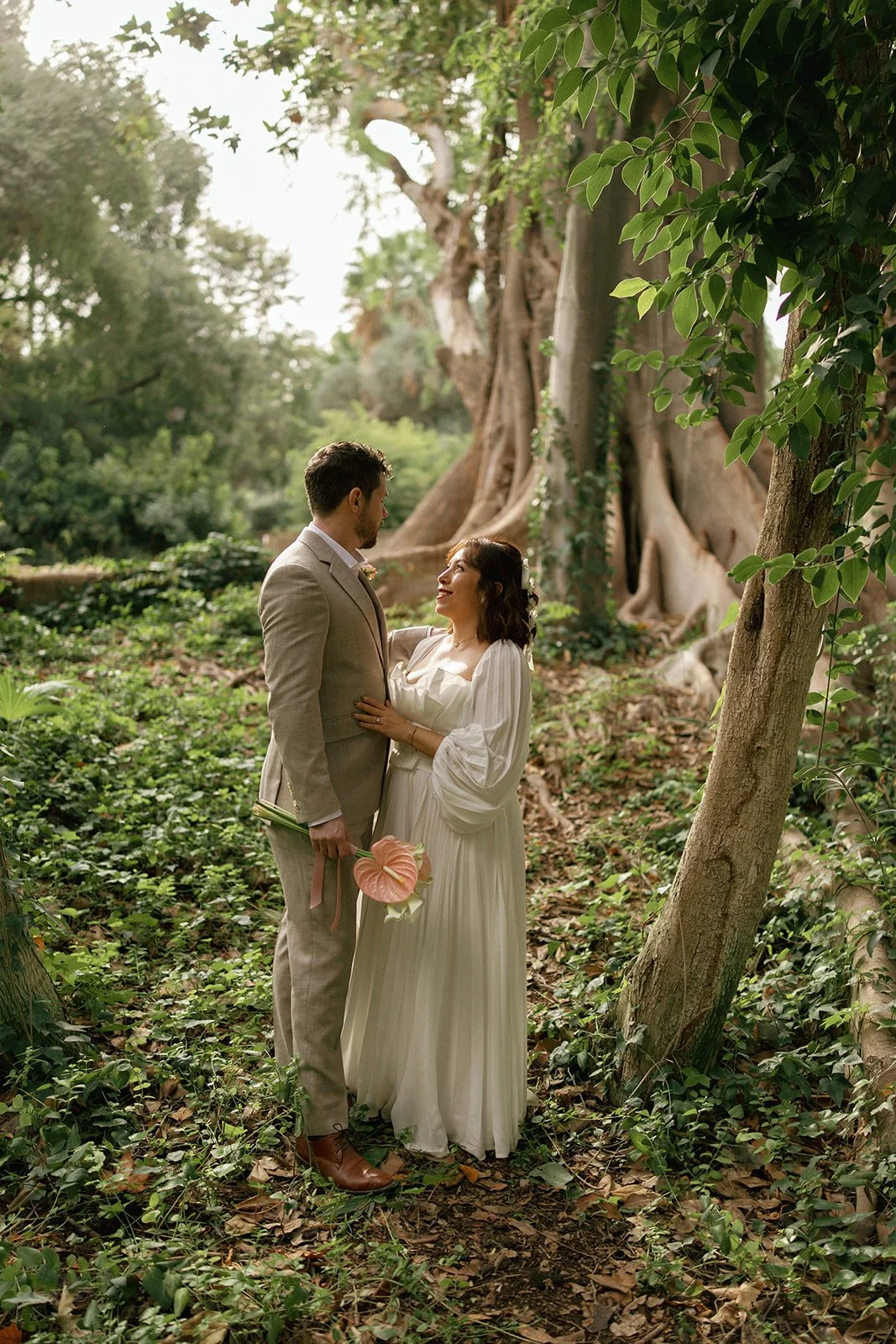 A couple in wedding attire stands close in a lush, green forest, gazing into each other's eyes, with a large tree in the background and soft sunlight filtering through the leaves.