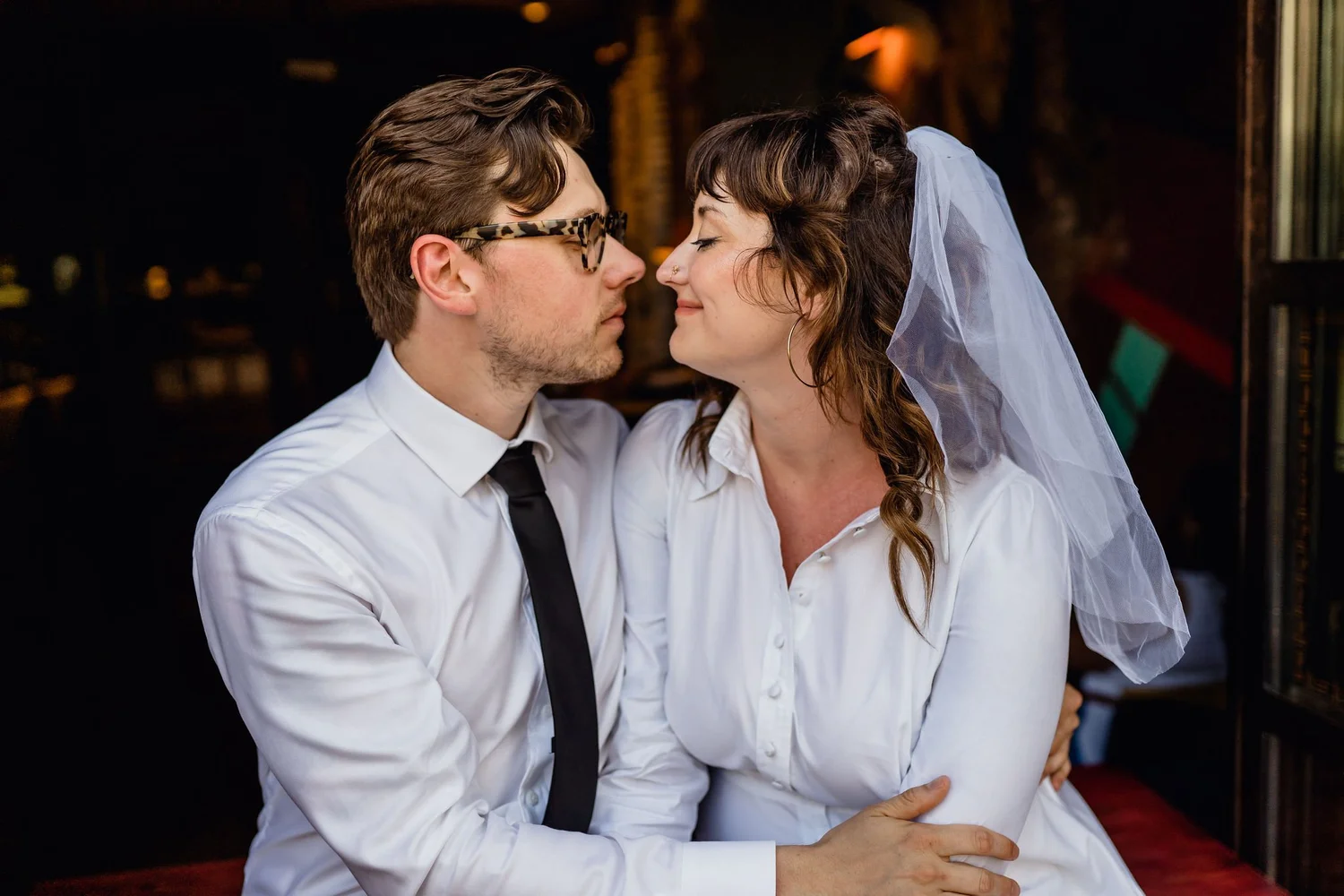 A couple on their wedding day, embracing closely with their foreheads touching and eyes closed, dressed in white shirts with the woman wearing a veil.