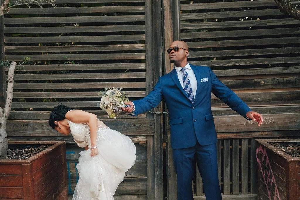 A man in a blue suit holding a bouquet funnily pulling a woman in a wedding dress by a chain as she bends over.
