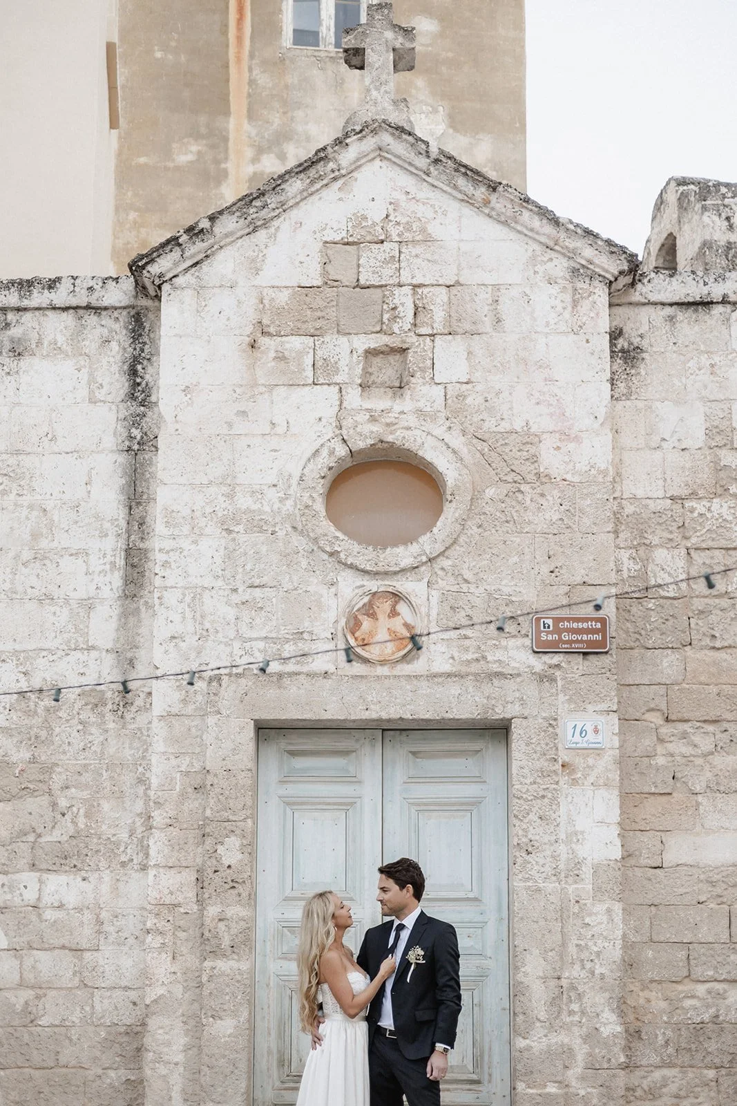 A couple in formal attire, a woman with a white dress and a man in a suit, are standing close to each other in front of an old stone building with a large, closed wooden door and religious symbols at the top.