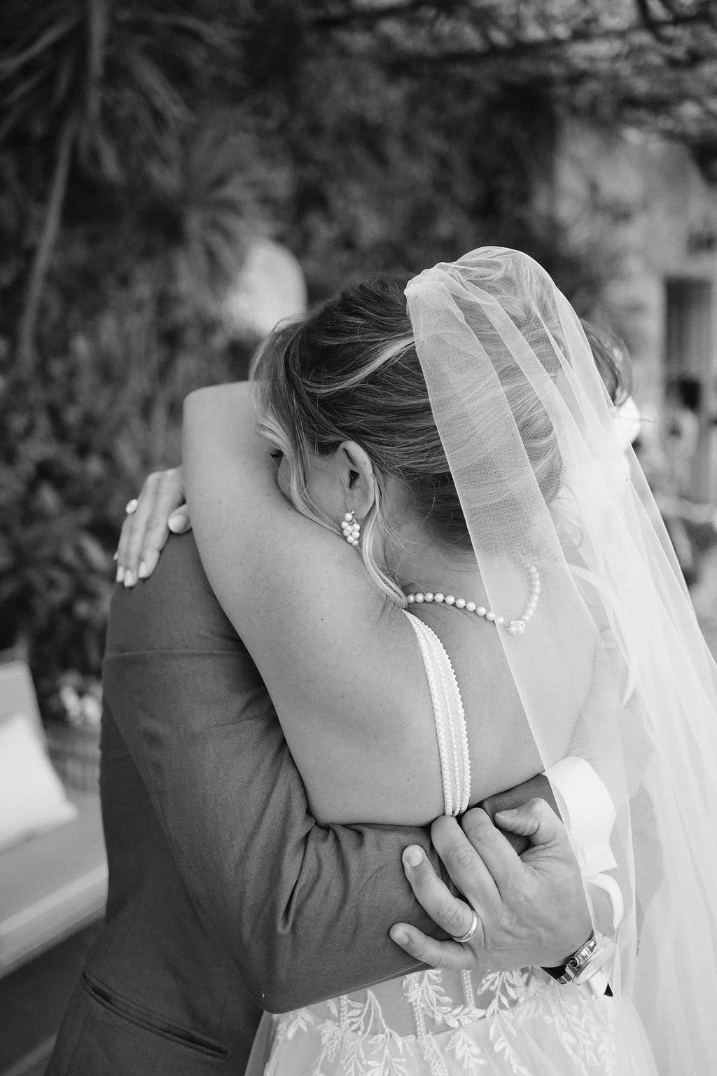 A bride embraces a person, with her veil and pearl jewelry visible, in an outdoor setting.