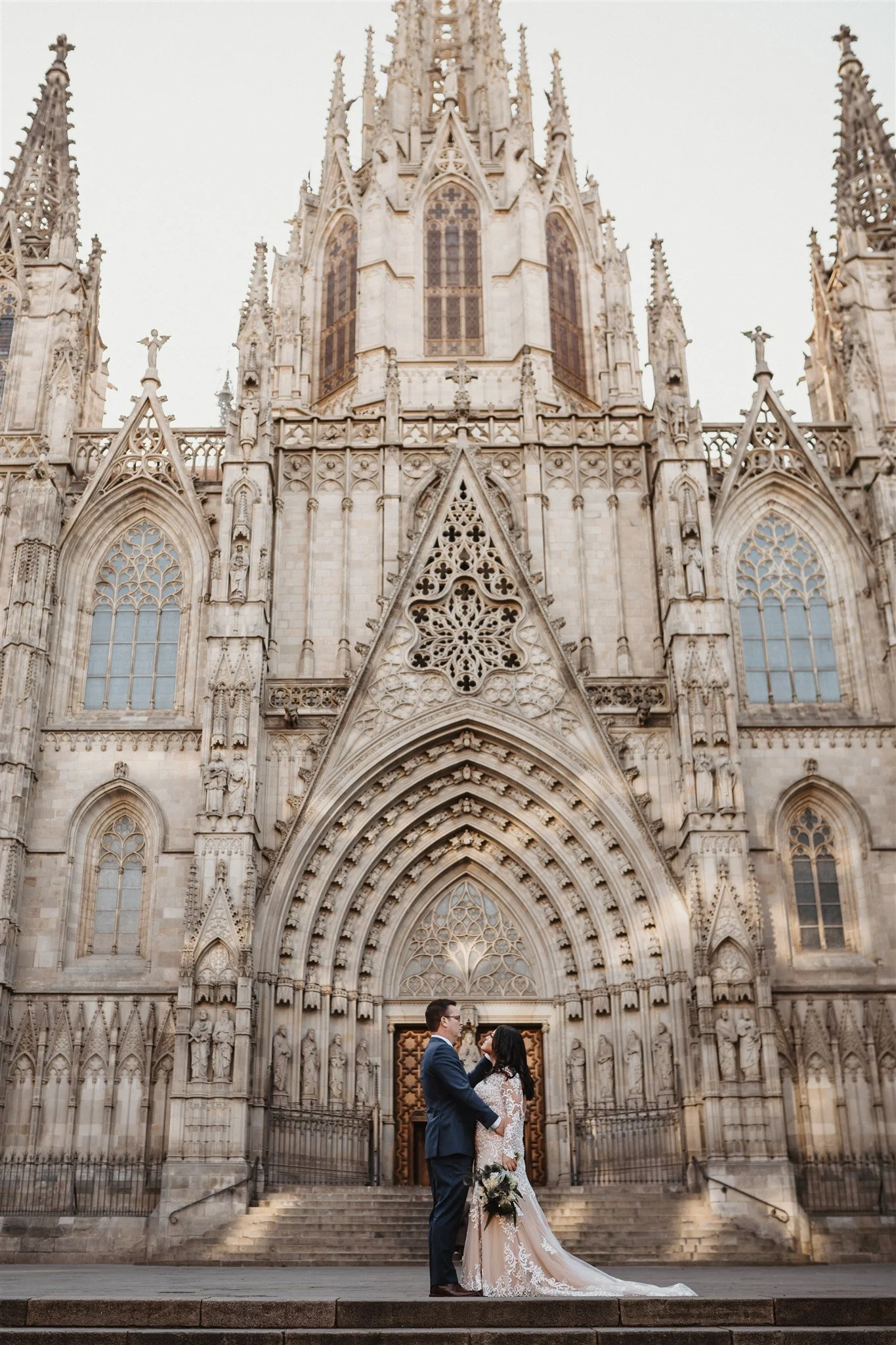 A bride and groom standing in front of a gothic cathedral, gazing into each other's eyes. The bride is wearing a lace wedding gown and holding a bouquet, while the groom is dressed in a dark suit. The architecture of the cathedral features intricate stone carvings and pointed arches.