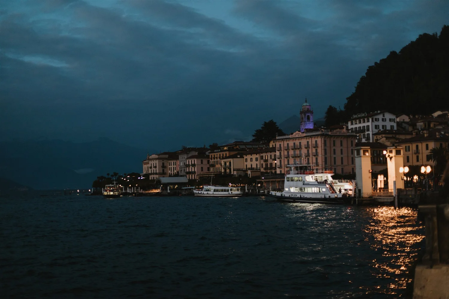 A coastal town at dusk with boats docked at the harbor, illuminated buildings, and a hill with a church tower in the background under a cloudy sky.