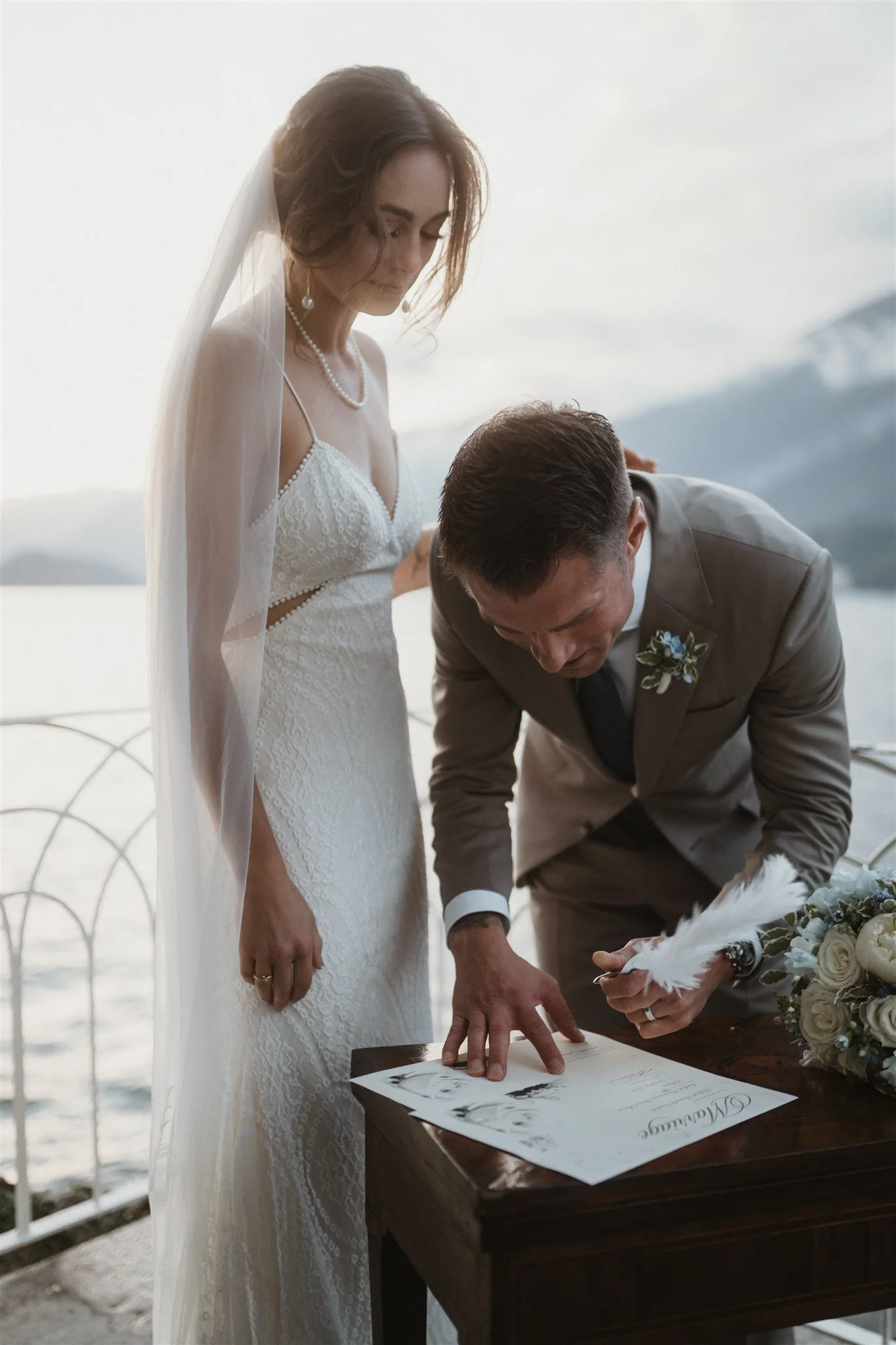 A bride and groom signing their marriage certificate outdoors with a body of water and mountains in the background.