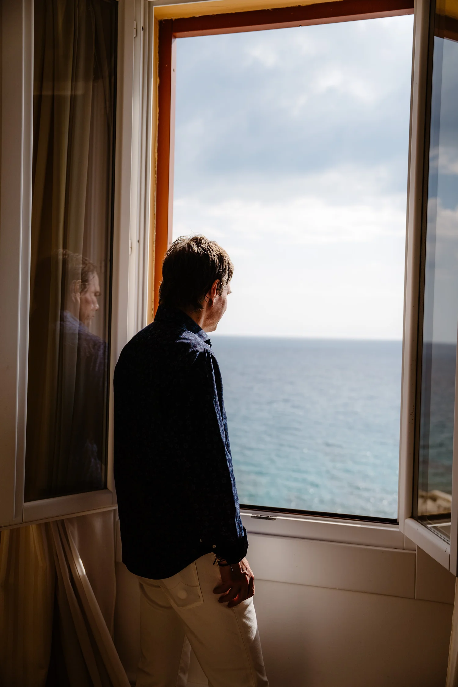 A person standing by an open window looking out at the ocean, with their reflection visible on the left side of the window.