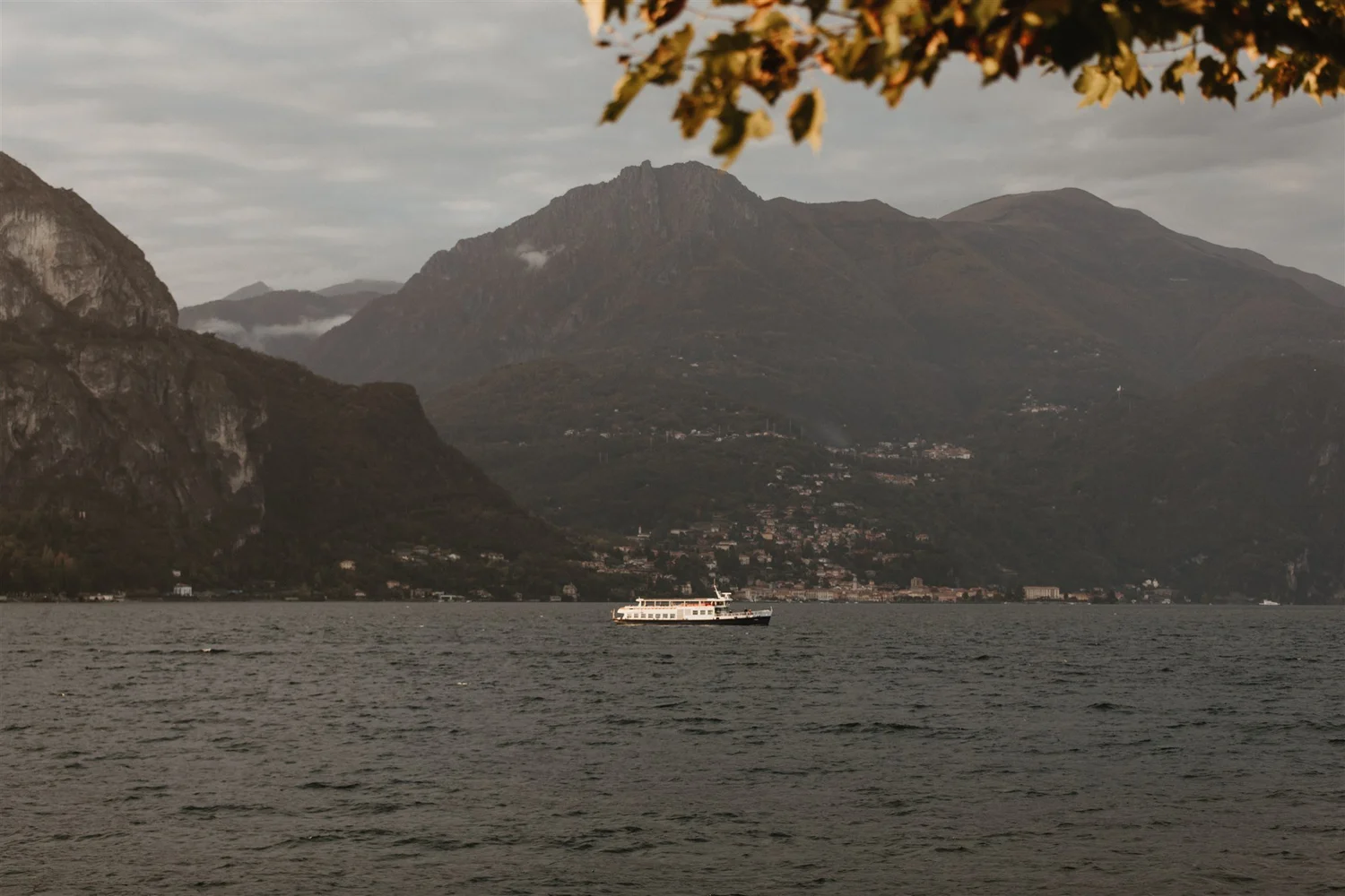 A large body of water with a passenger ferry boat in the foreground. In the background, there are tall, rugged mountains with a cloudy sky above.