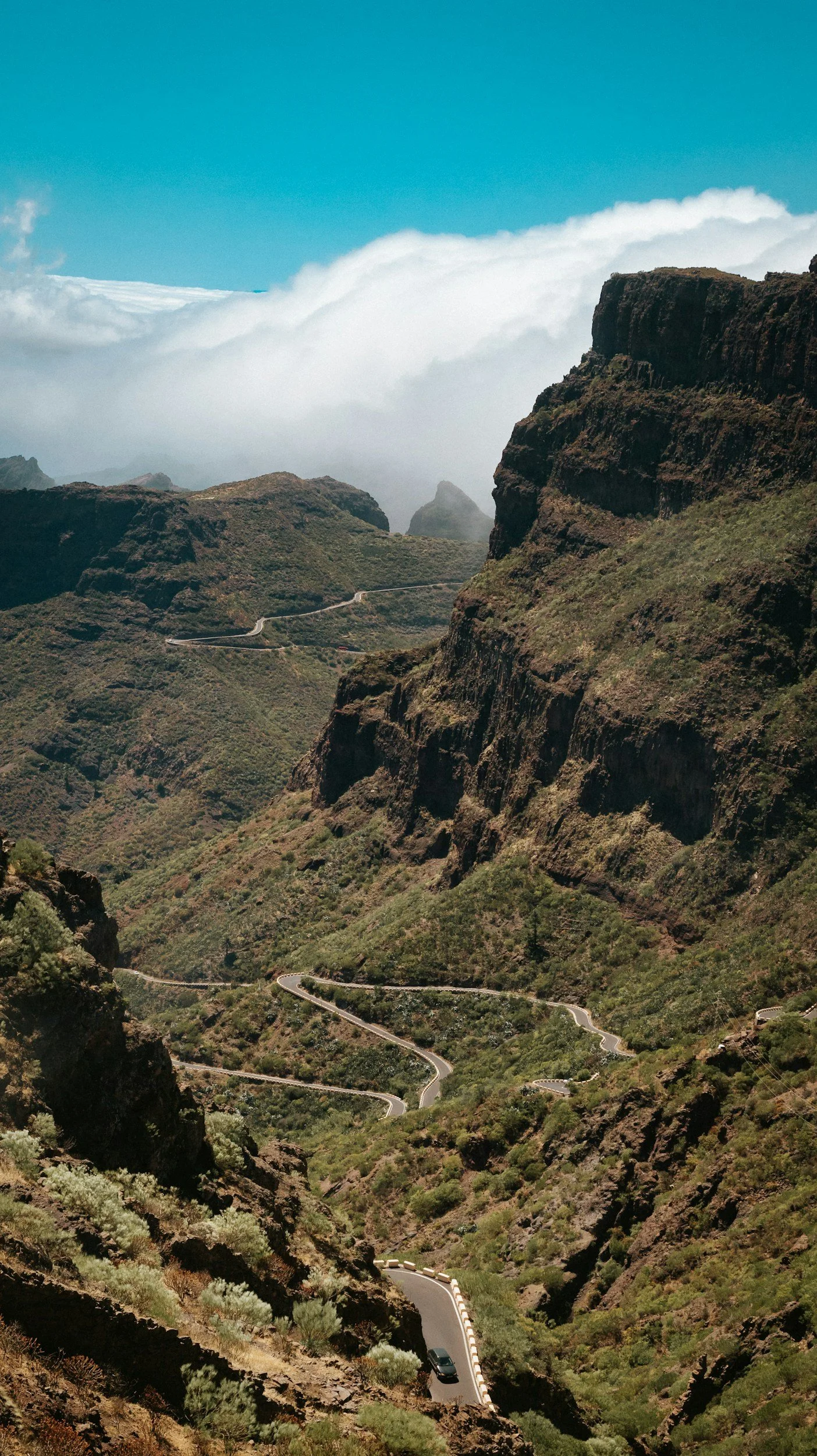 A winding mountain road through a green canyon with steep cliffs and cloudy sky.