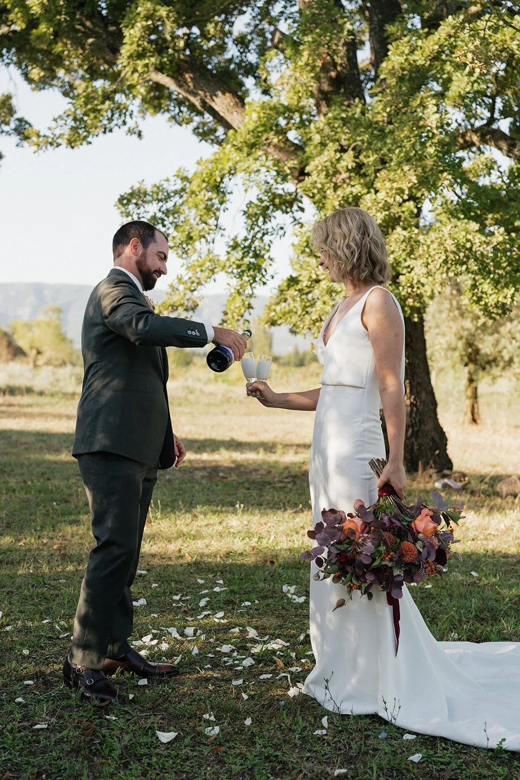 A couple at their outdoor wedding, with the groom pouring champagne into a flute held by the bride, who is holding a large bouquet of flowers.
