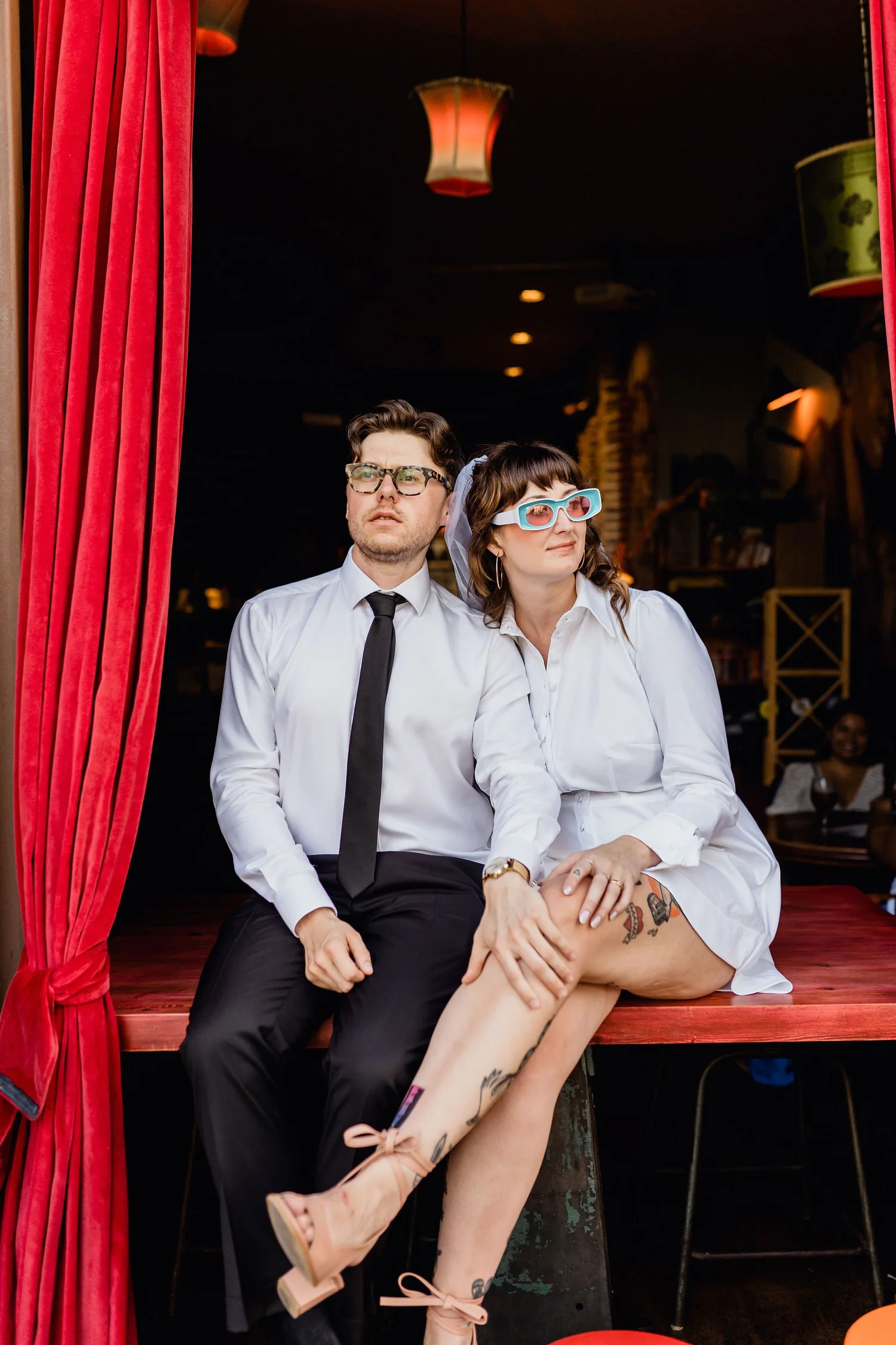A man and a woman sitting together on a stage with red curtains behind them. The man has glasses, a white shirt, and a tie. The woman is wearing glasses, a white shirt, and shorts, with tattoos on her legs. The woman is leaning on the man with her le