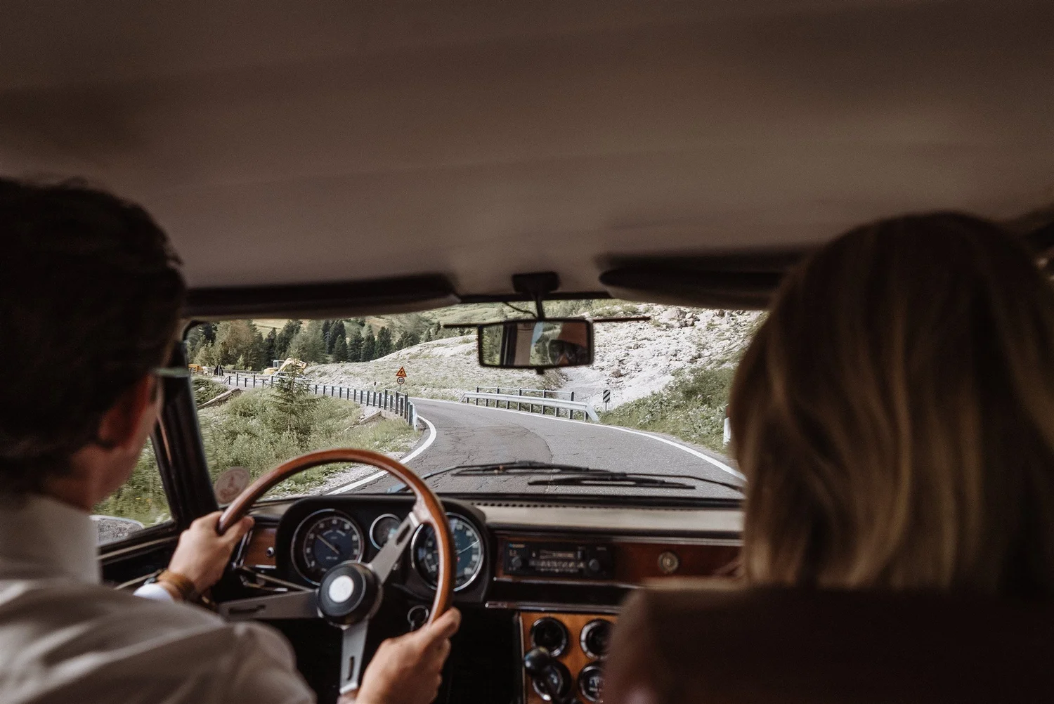 View from inside a car with two people, one driving and one in the passenger seat, on a winding road through a rocky and forested landscape with railings and road signs.