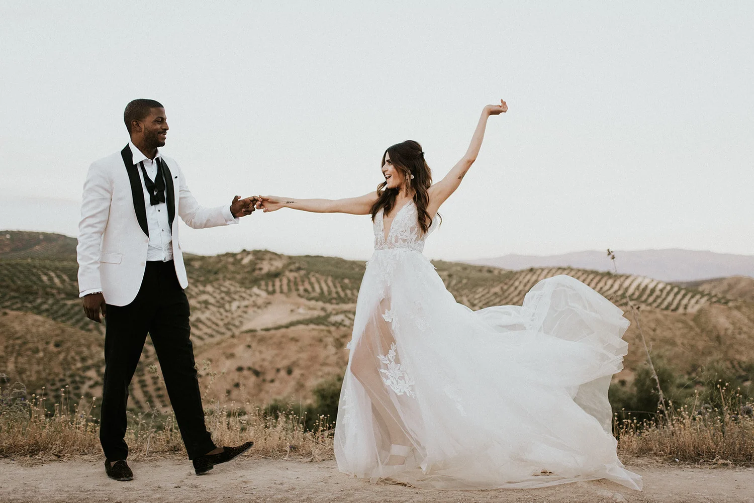 A couple in wedding attire dancing outdoors on a dirt road with rolling hills in the background. The woman is wearing a white wedding gown with floral lace details, and the man is dressed in a white blazer, black pants, and a black tie. He is holding