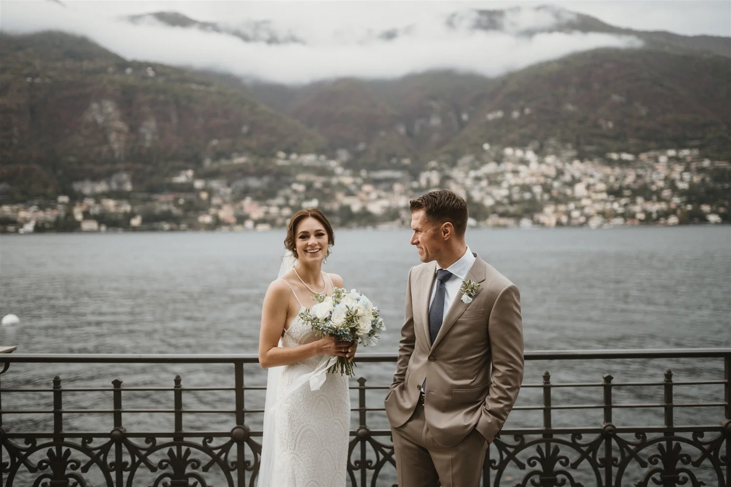 Bride and groom standing by a railing near a body of water with a picturesque mountainous landscape and cloudy sky in the background.