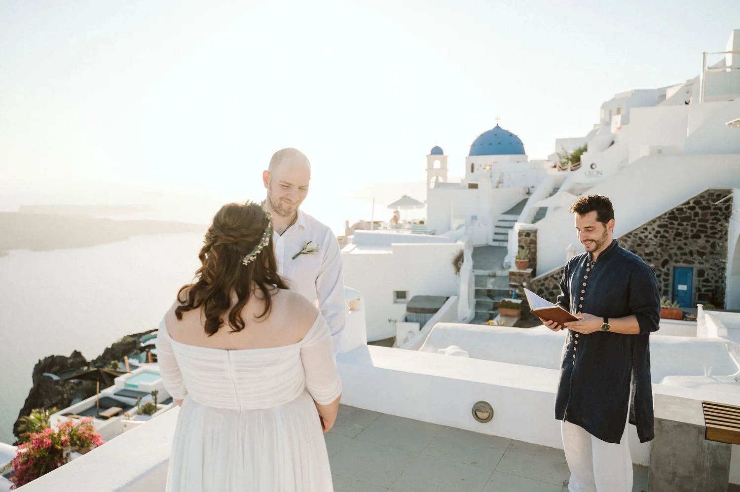 A wedding ceremony taking place outdoors in front of white buildings with blue domes, in Santorini, Greece. A bride with brown hair and a white off-shoulder dress is facing the groom, with an officiant reading from a book.