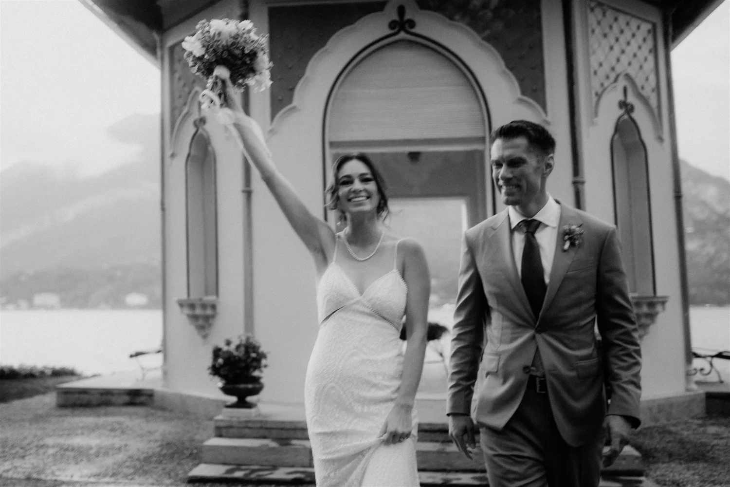 A black and white photo of a smiling woman in a white wedding dress holding a bouquet and a man in a suit holding her hand, as they walk together, probably on their wedding day.
