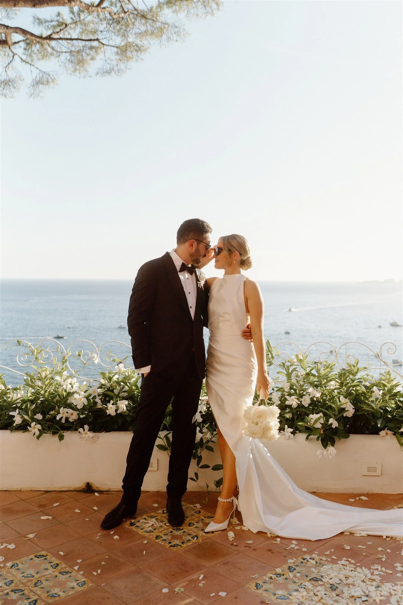 A bride and groom standing close together by a decorated outdoor ledge, holding hands and touching foreheads, with an ocean view in the background during daytime.