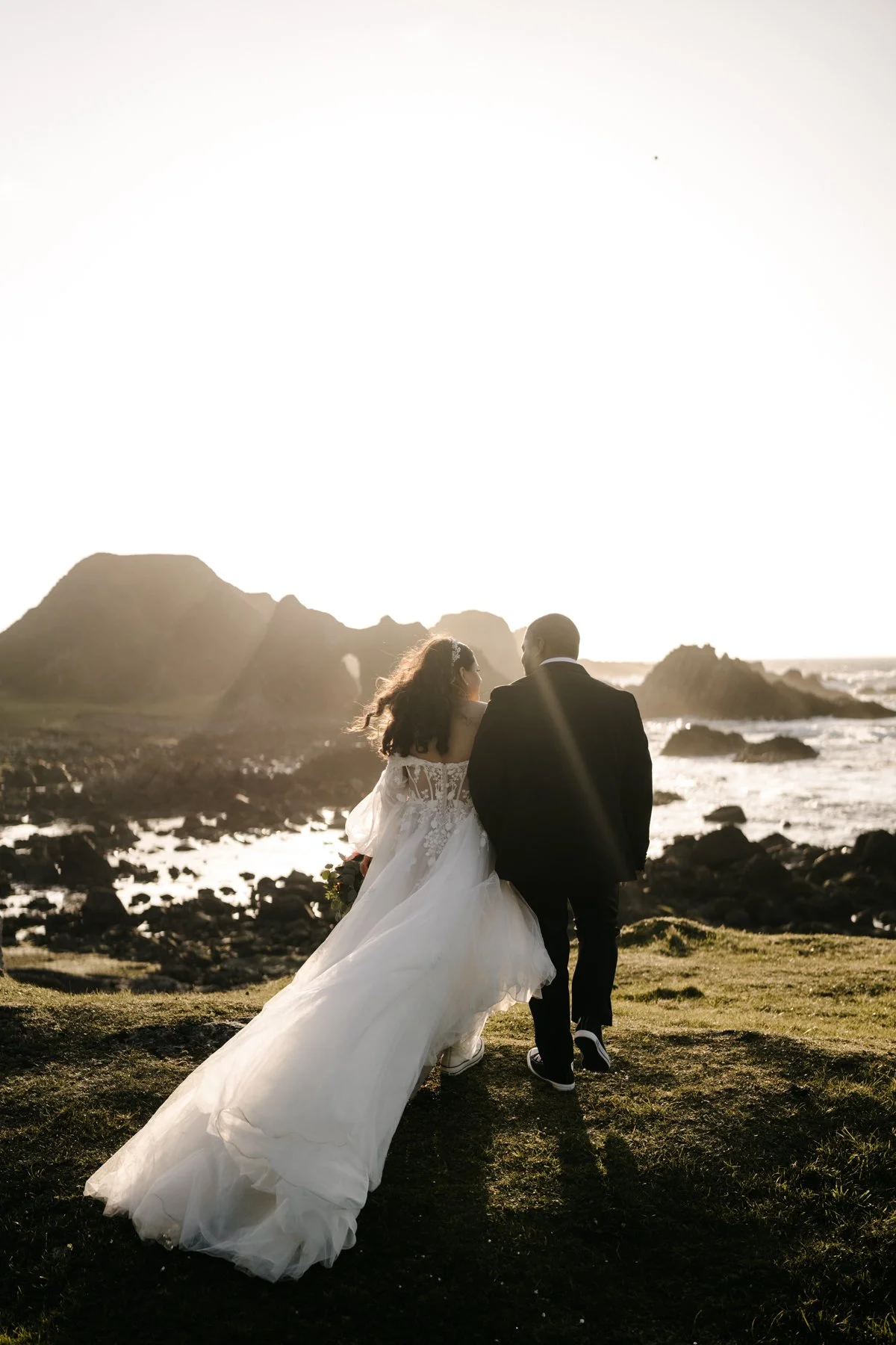 A bride and groom walking along a grassy beach at sunset, with rocky formations and the ocean in the background.