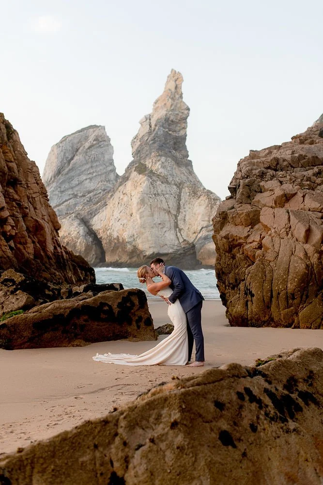 A couple in wedding attire kissing on a beach at sunset, surrounded by large rocks and a prominent rock formation in the background.