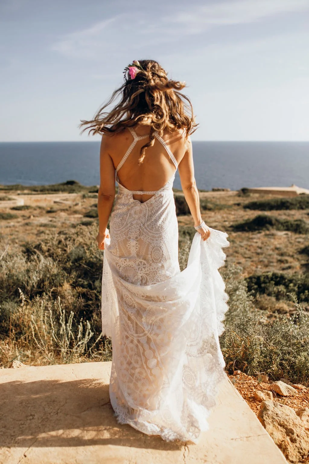 A woman in a white lace dress walking along a rocky path with the ocean in the background on a sunny day.