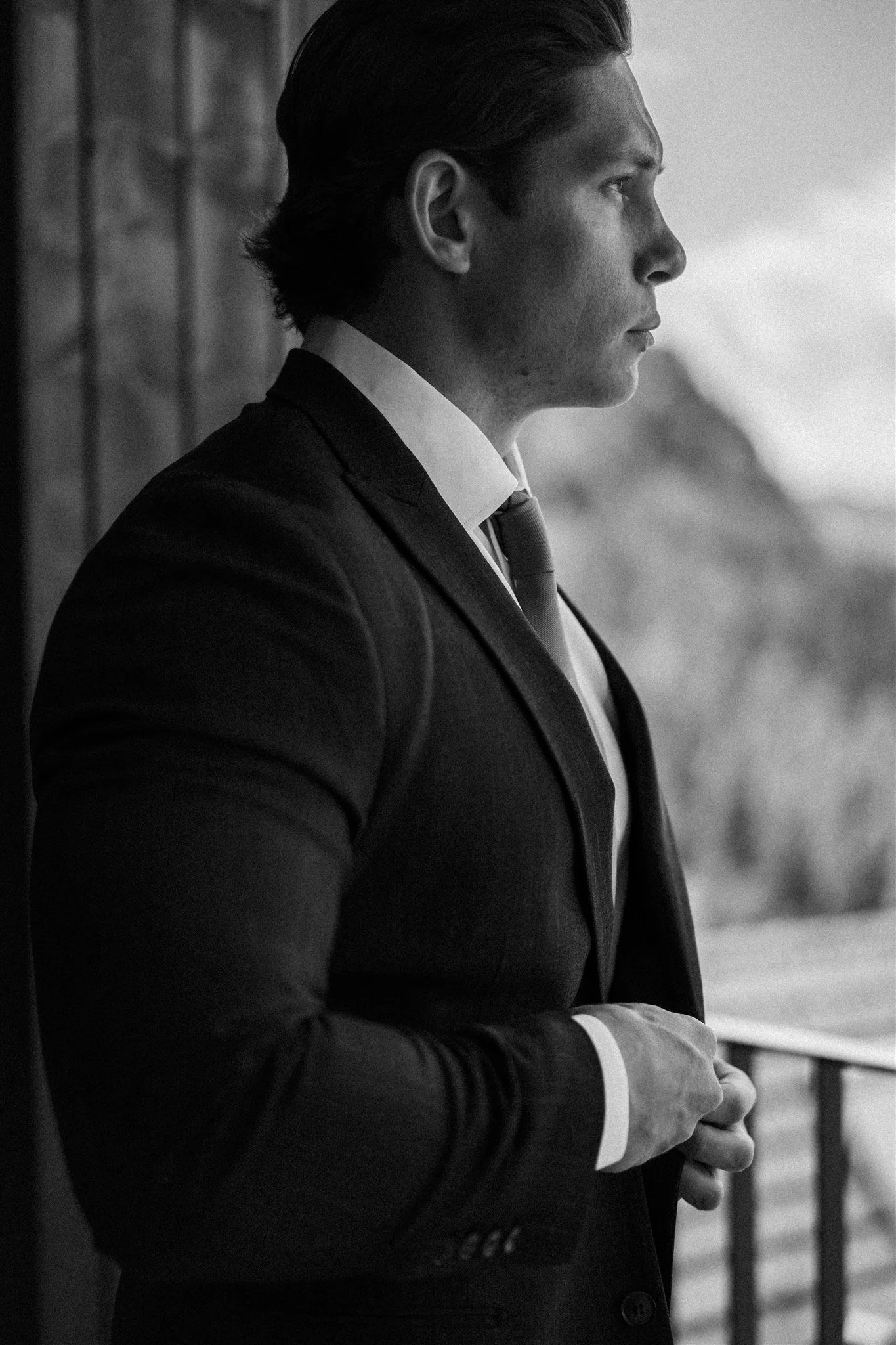 A man in a suit with a tie, standing outdoors, looking to the right, with a railing and blurred mountainous landscape in the background, captured in black and white.