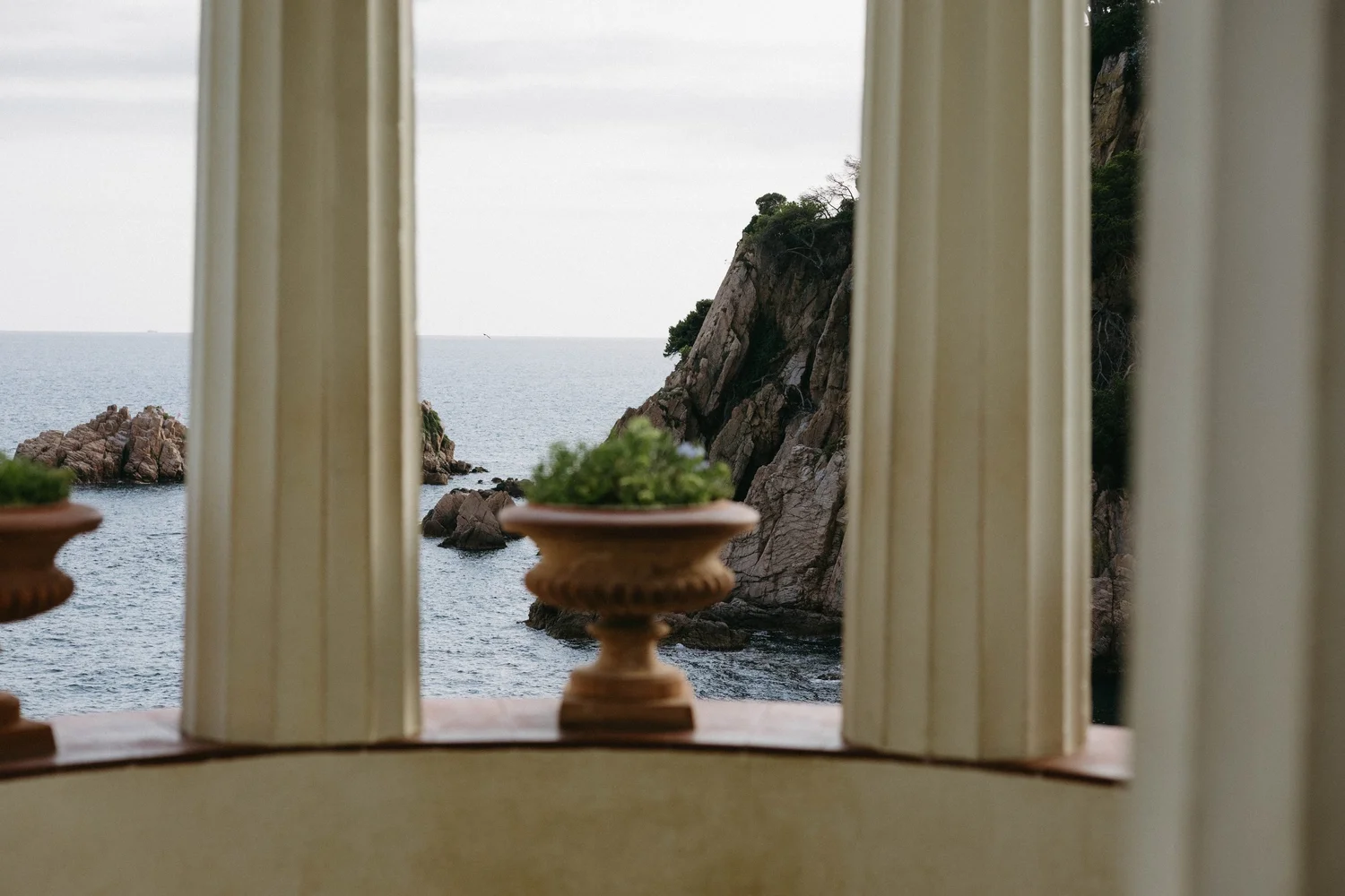 View of rocky coastline and the ocean through a window with beige curtains, with two small potted plants on a window sill.