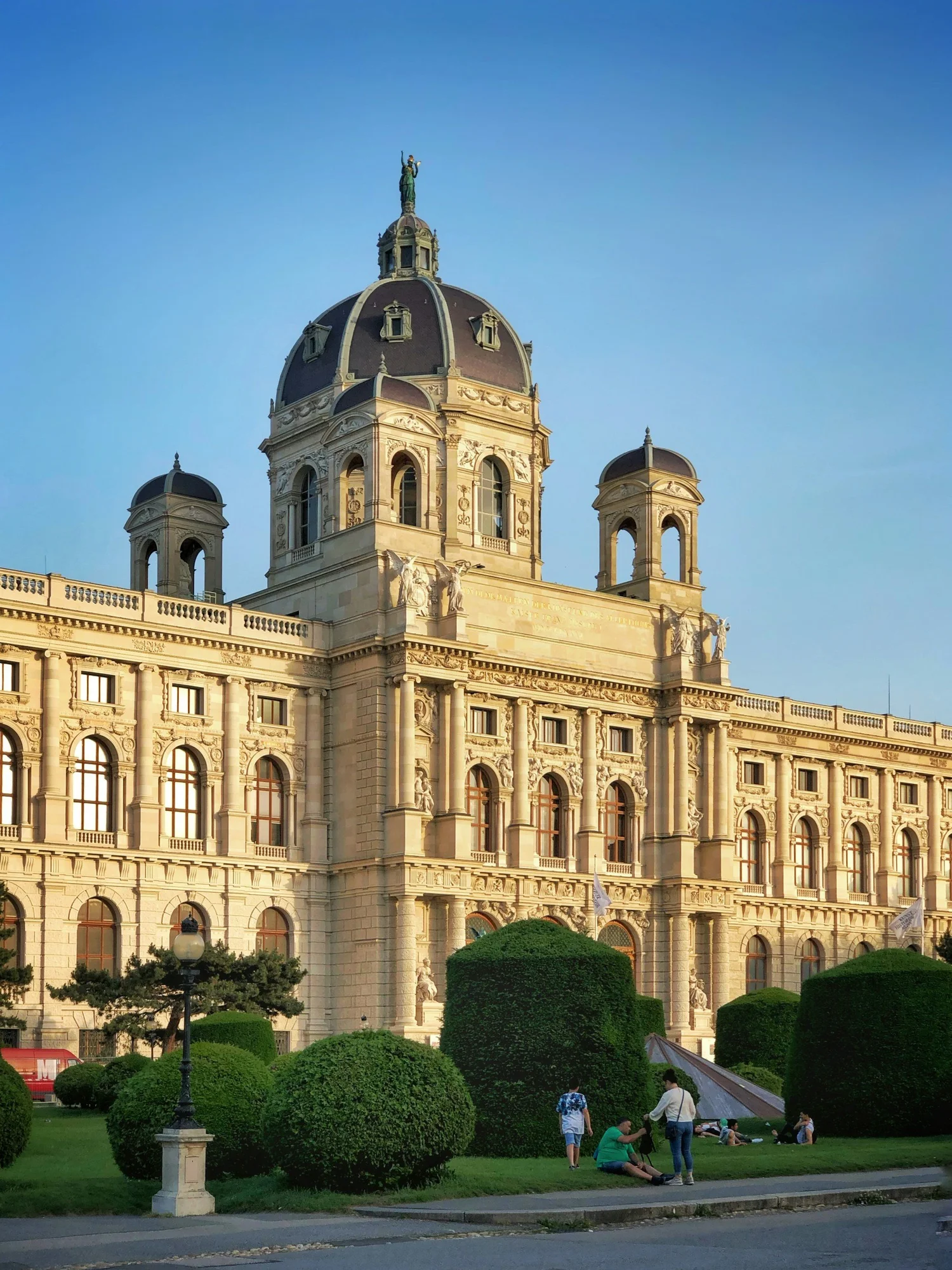 A historic building with ornate architecture and a dome topped with a statue, surrounded by a well-maintained park with trimmed bushes and people relaxing and walking.