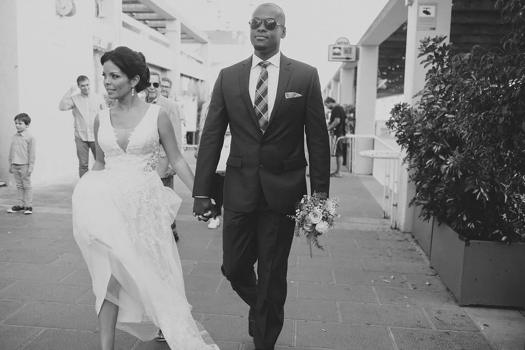 A bride and groom walking outdoors, with the bride in a lace wedding dress and the groom in a dark suit, holding a small bouquet of flowers, surrounded by onlookers.