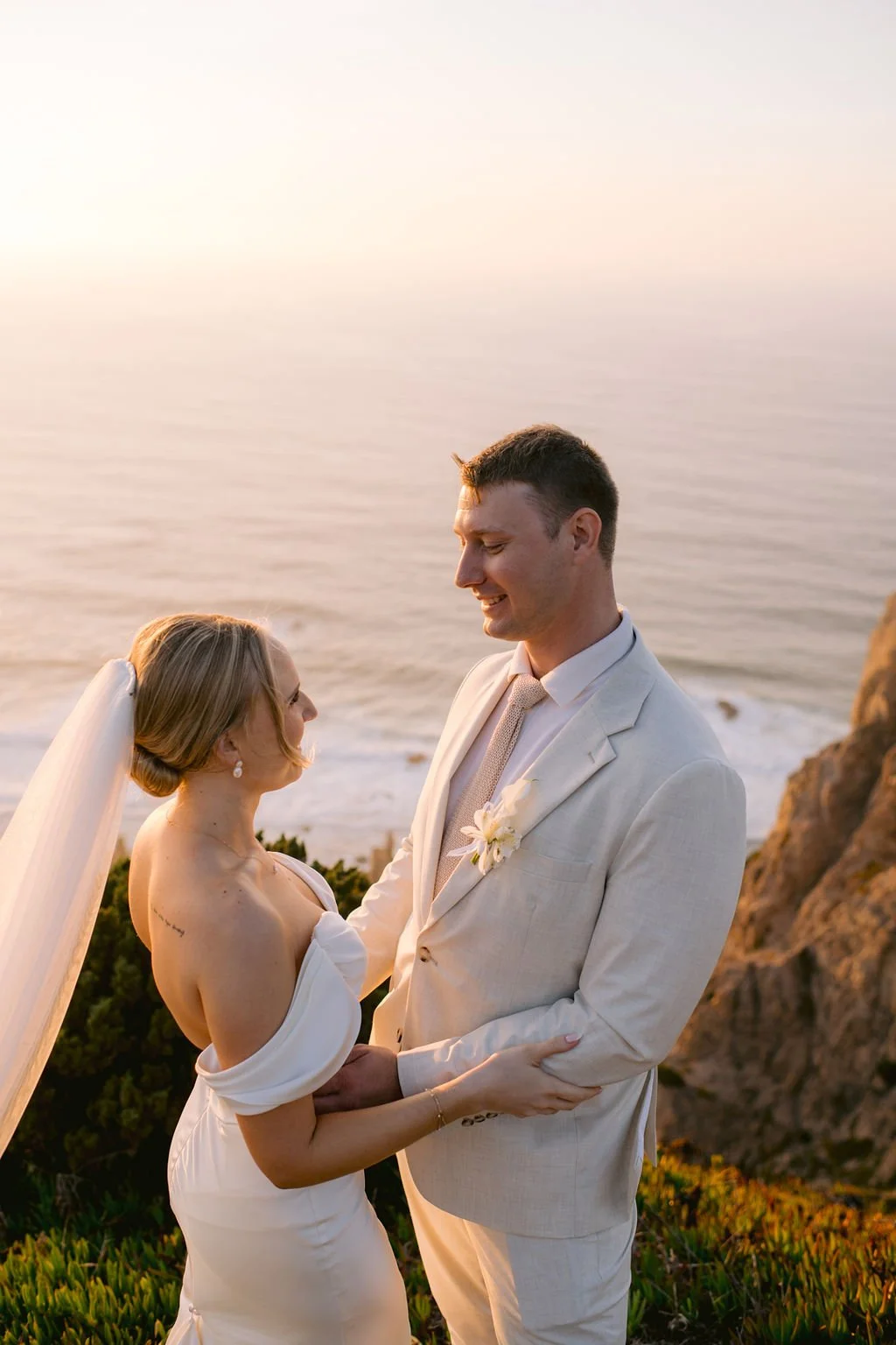A bride and groom standing close together on a cliffside overlooking the ocean at sunset, smiling at each other during their wedding.